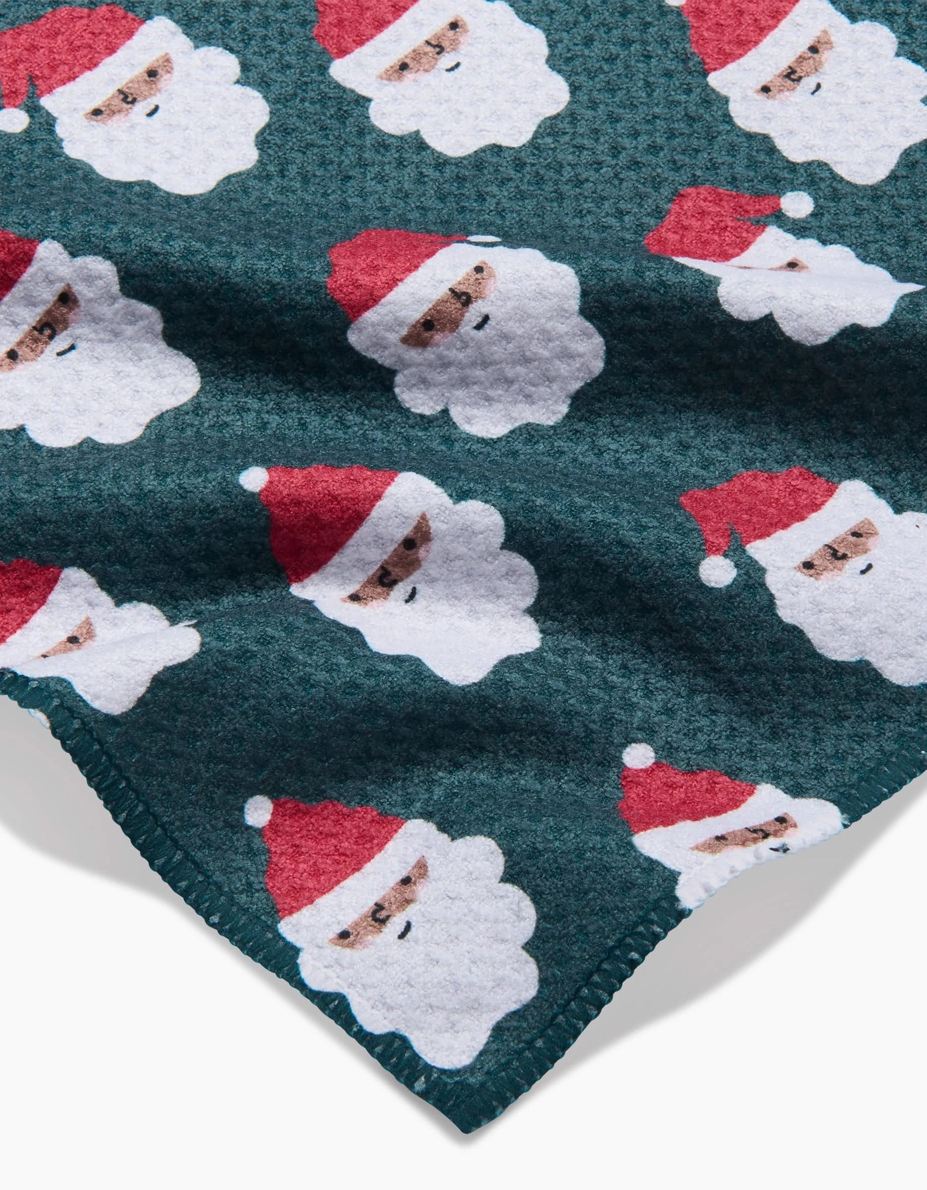 The Santa Santa towel by Charlie Rowan Designs is dark green, ultra-absorbent, and textured with a repeating pattern of cartoon Santa Claus faces in red hats and white beards. The towel’s edge appears in the lower corner.