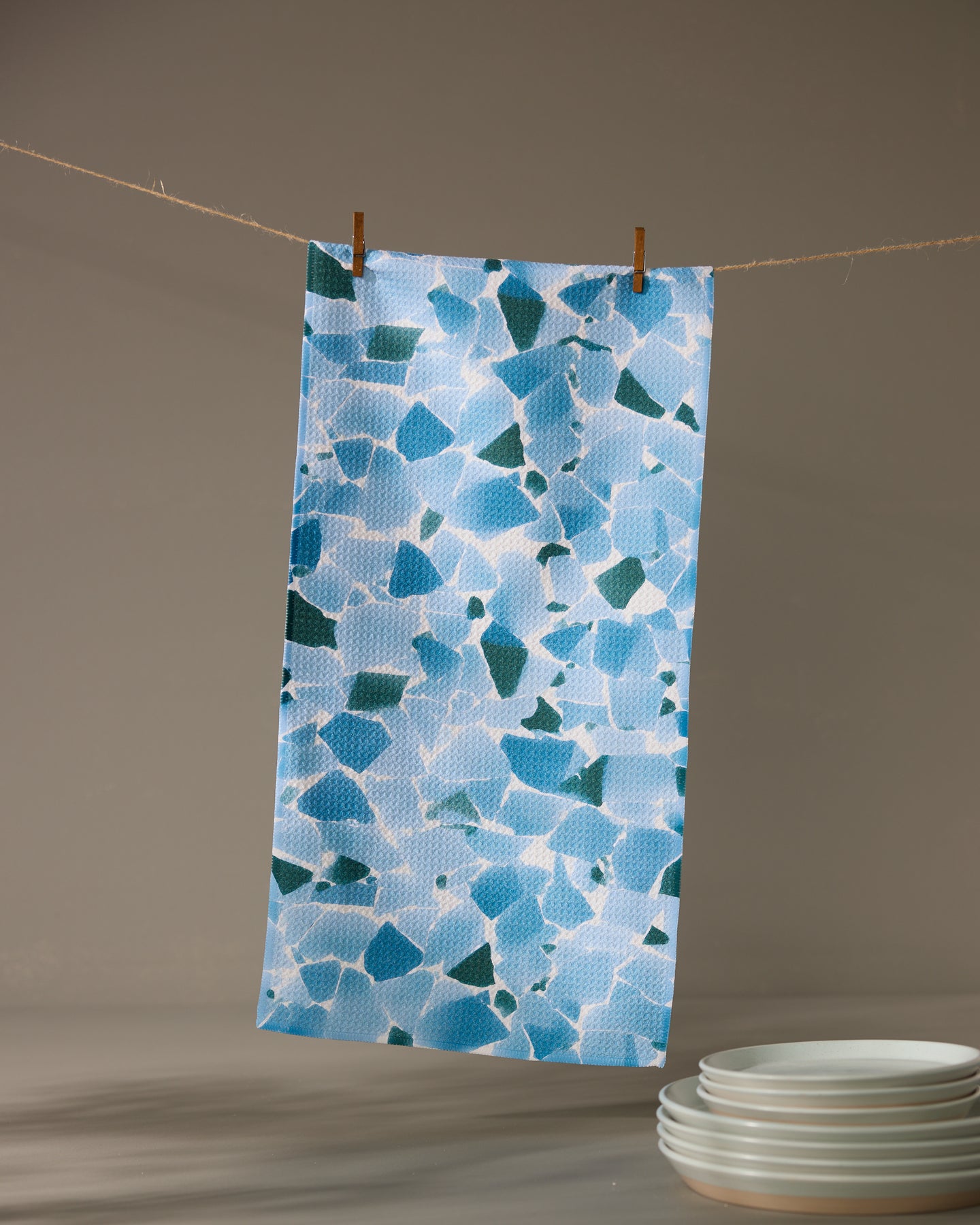 The Geometry Seaglass ultra-absorbent towel in a blue and white pattern hangs from a clothesline with wooden clothespins, above a stack of white plates on a gray surface.