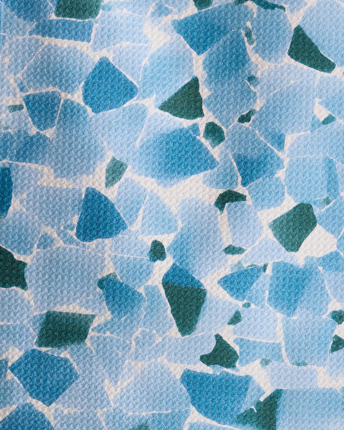 Seaglass by Geometry features an abstract mosaic of irregular blue and green shapes on a textured white background, evoking recycled materials. The random arrangement creates an artistic, modern design.