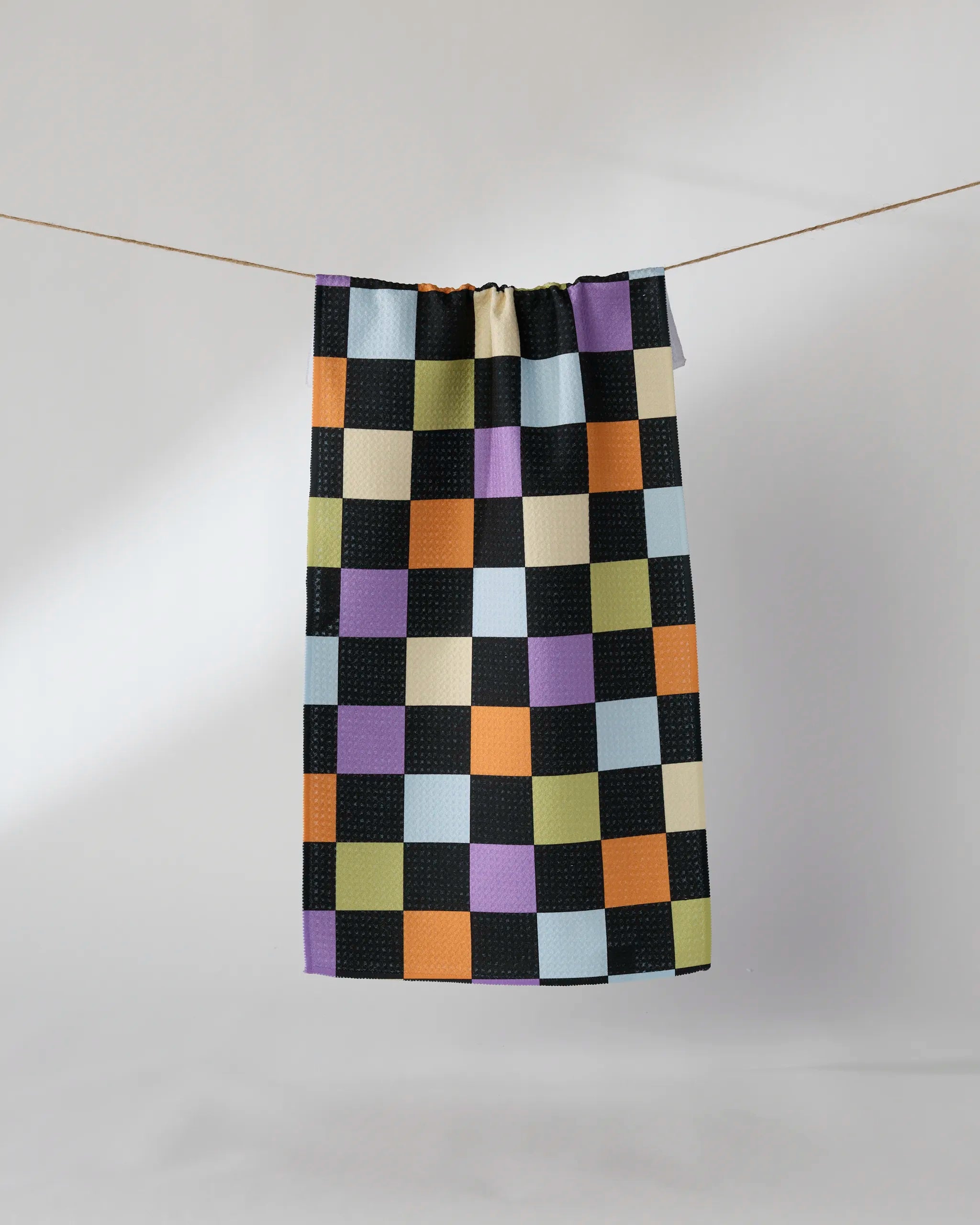 The Geometry See You Checkers towel, featuring ultra-absorbent fabric and a black, purple, orange, yellow, green, and light blue checkered pattern, hangs on a clothesline against a plain light background.