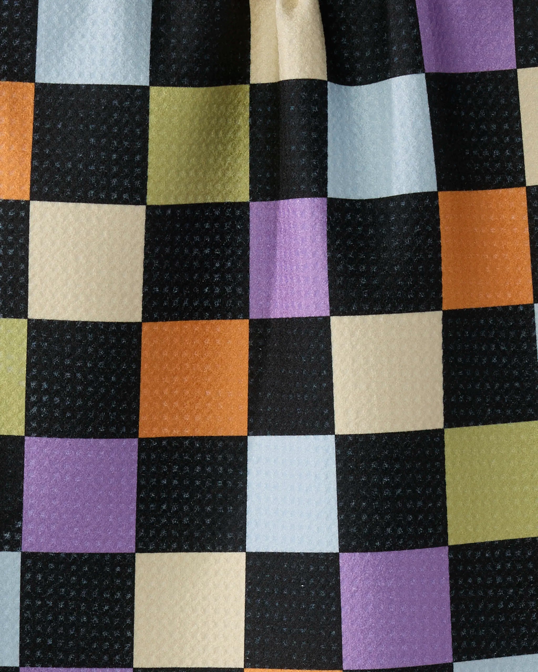 A close-up of Geometry’s See You Checkers recycled kitchen towel shows black, cream, orange, green, purple, and light blue squares with small textured dots. This ultra-absorbent towel features slight folds for added dimension.
