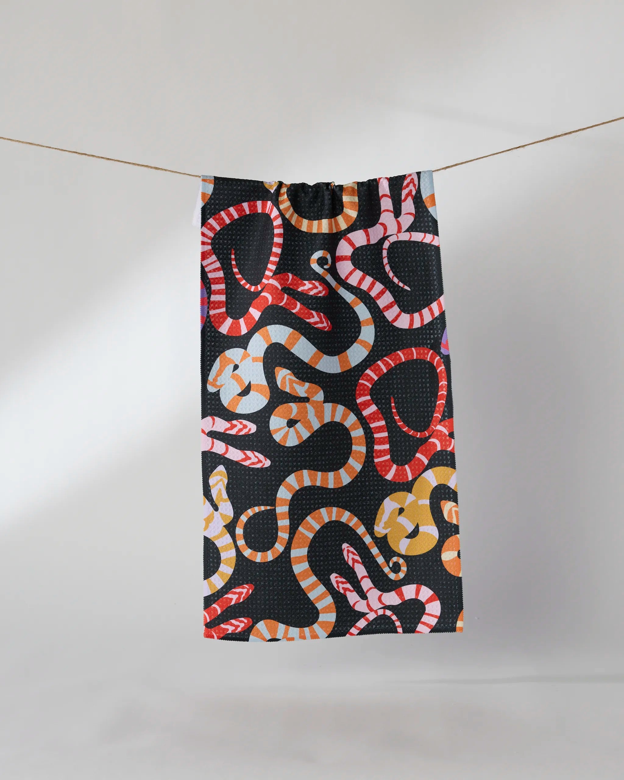 The Geometry Serpentine Riot recycled kitchen towel, featuring a bold snake pattern in red, orange, yellow, white, and pink, hangs on a clothesline against a plain white background.