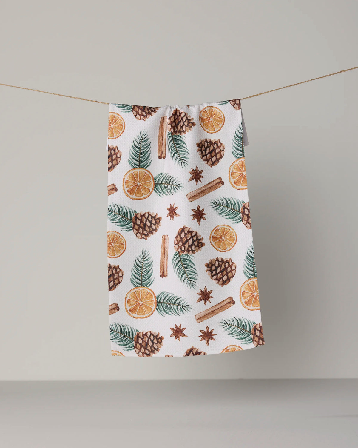 A Geometry Simmer Pot recycled kitchen towel in white, featuring a festive pattern of pinecones, orange slices, green leaves, cinnamon sticks, and star anise, hangs on a string against a plain background.