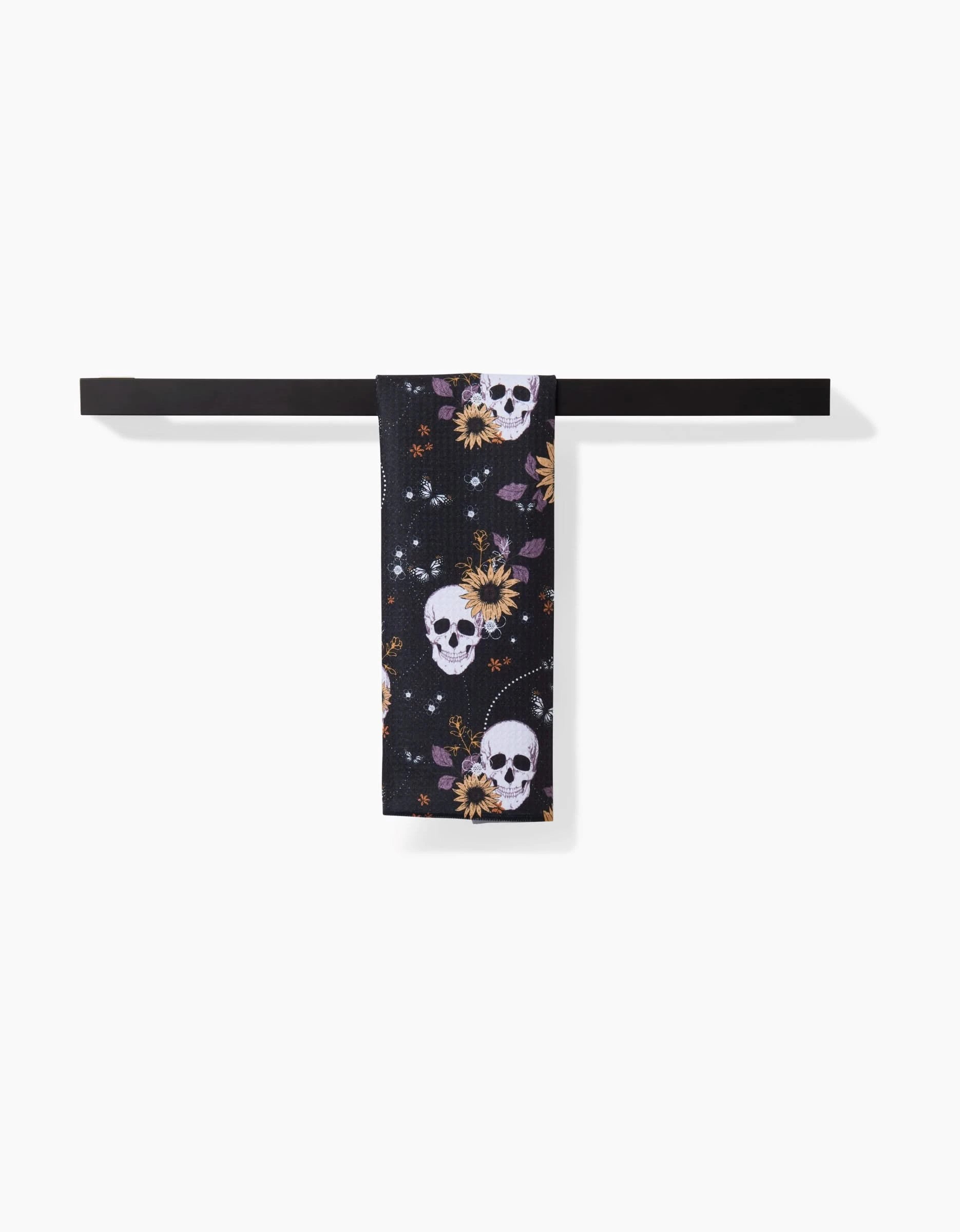 The Skulls In Bloom bar towel by Ashley Celena, featuring white skulls, yellow sunflowers, and small leaves, hangs neatly folded over a sleek black towel bar against a plain white background.