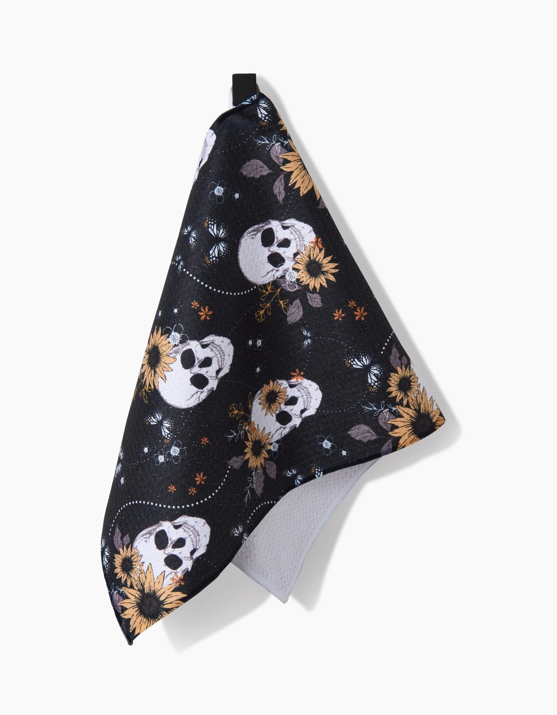 The Skulls In Bloom kitchen towel by Ashley Celena features black fabric with white skulls adorned with yellow sunflowers and blue flowers, shown hanging with a folded corner.