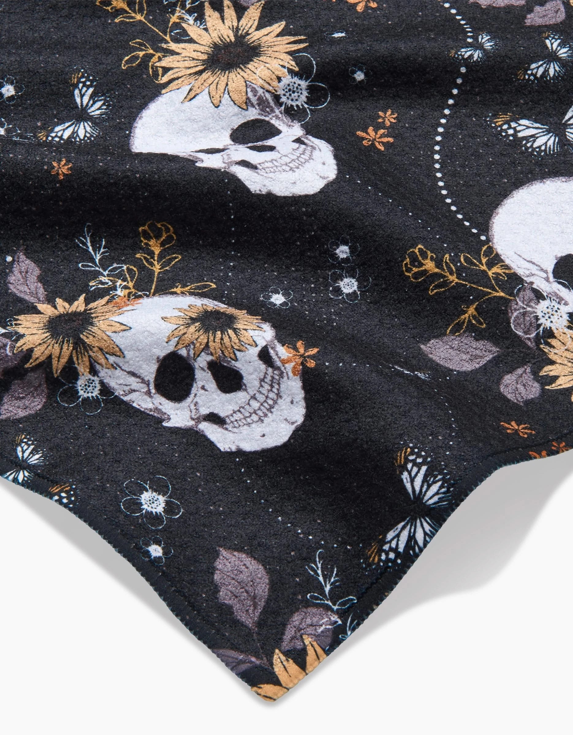 The Skulls In Bloom kitchen towel by Ashley Celena features a black fabric adorned with white skulls, yellow sunflowers, white butterflies, and gold botanical accents.