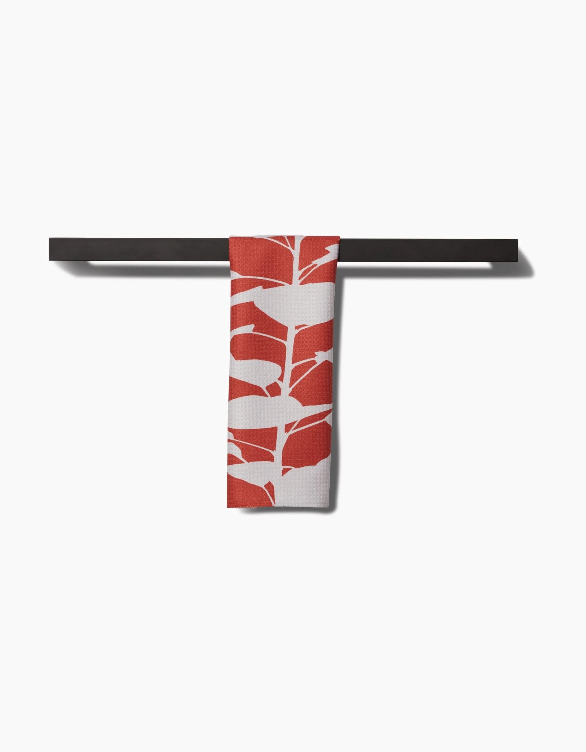 The PrintedPlant Smoketree bar towel, featuring red and white abstract leaf patterns on Geoweave fabric, hangs neatly over a minimalist black horizontal bar against a plain white background.