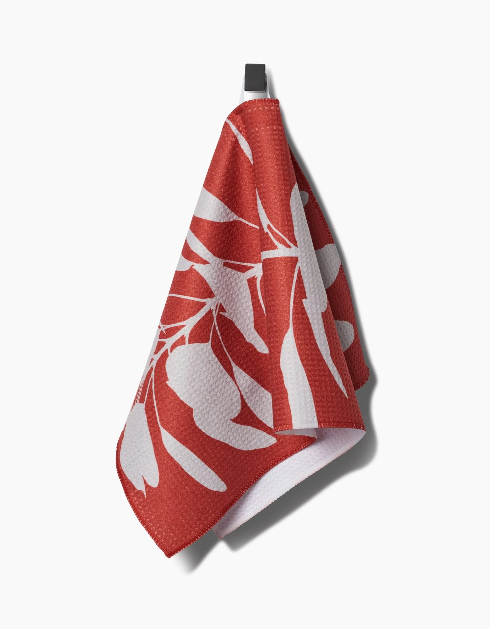 A PrintedPlant Smoketree bar towel, featuring white leaf patterns on red textured Geoweave fabric, hangs from a black loop against a white background. The towel is neatly draped, casting a soft shadow.