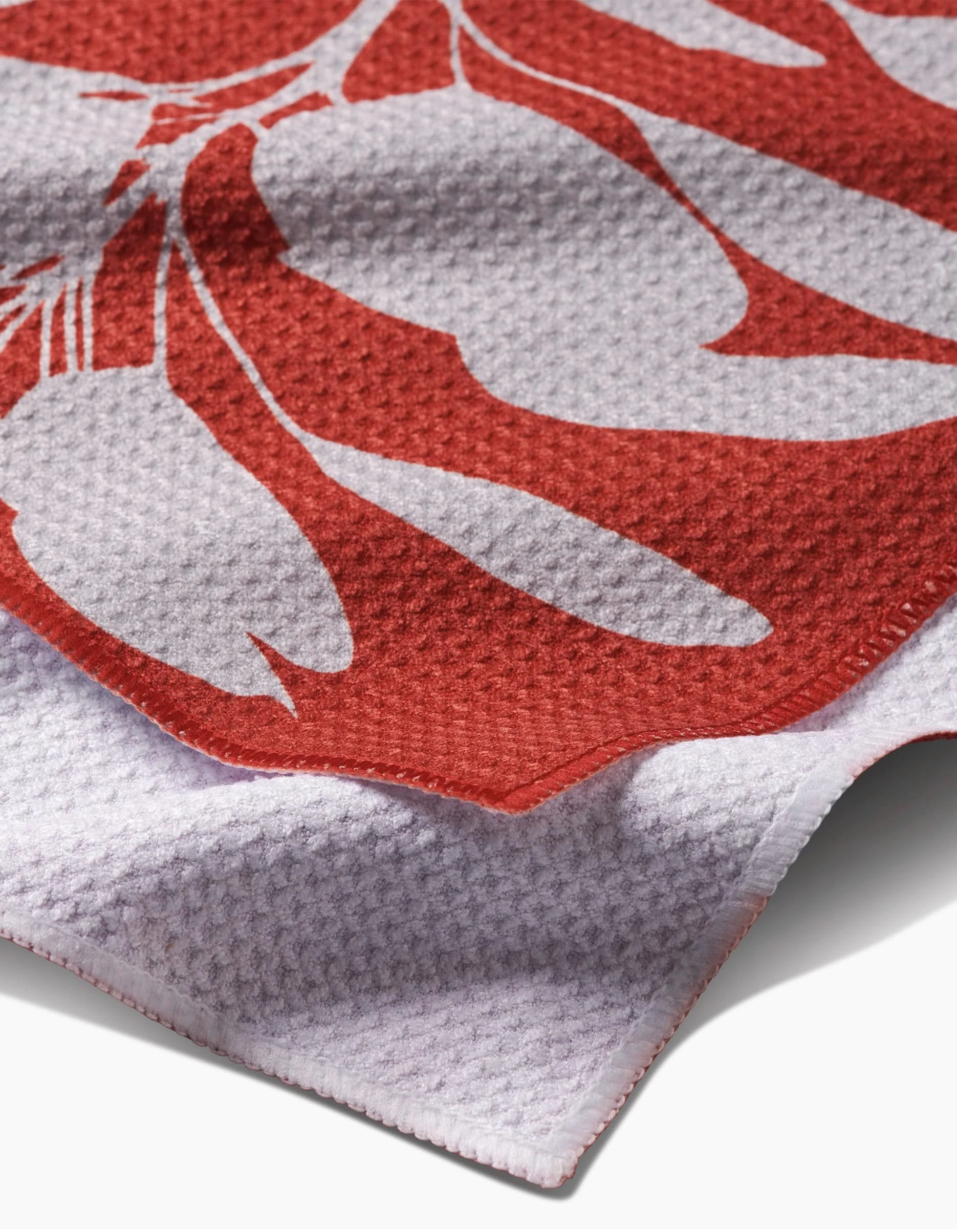 A close-up of the PrintedPlant Smoketree Bar Towel, crafted from Geoweave fabric, shows its red and white botanical pattern, textured weave, and an upward-folded edge.