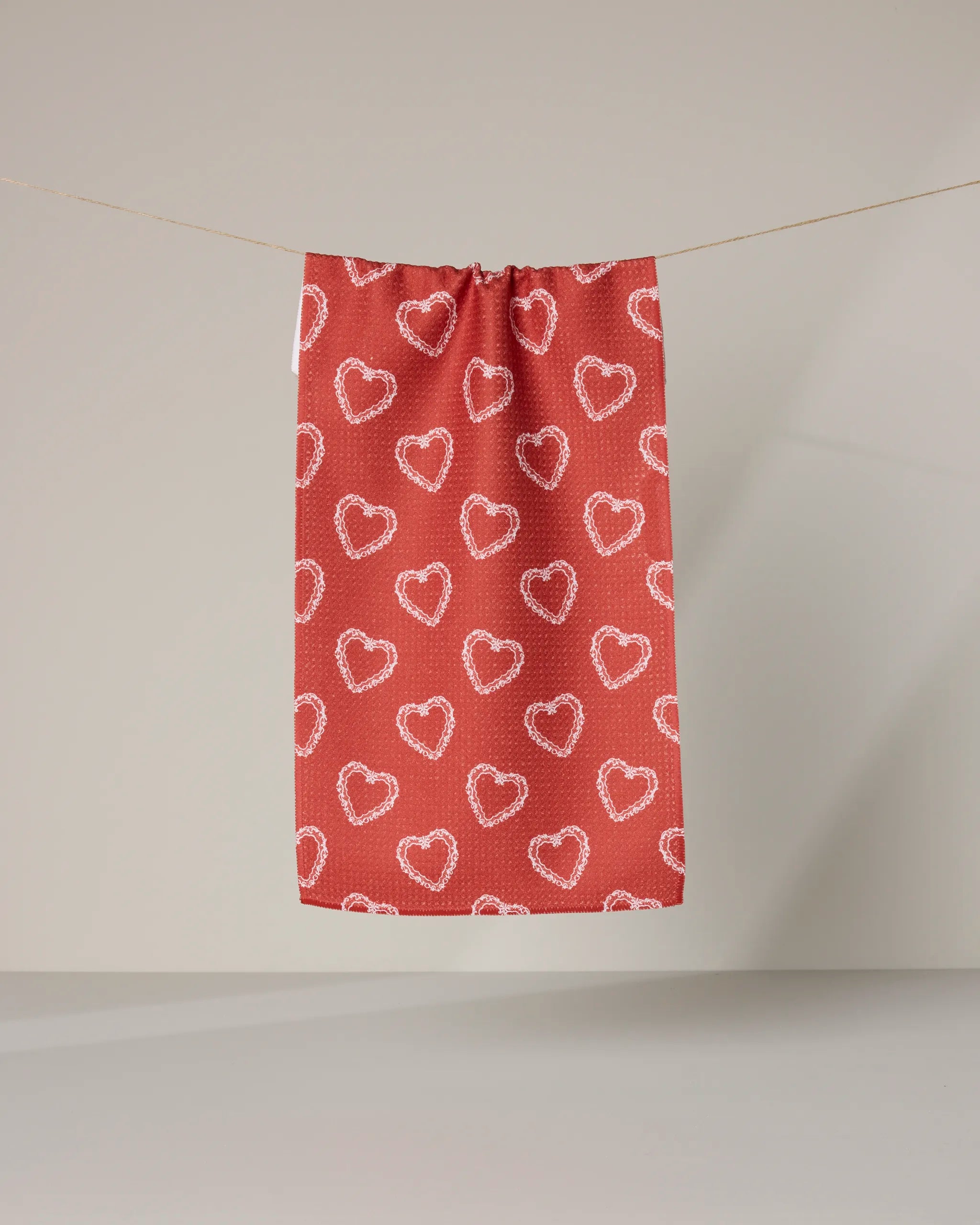 The Geometry "Soft Heart Scarlet" bar towel in red, with a white stitched heart outline pattern, hangs on a string against a plain, light-colored background.