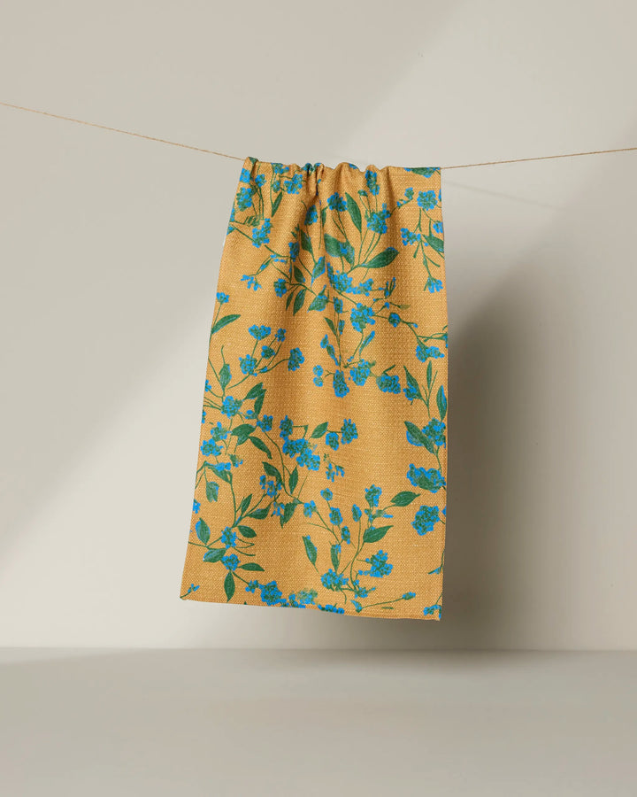 The Geometry Soleado towel, featuring a blue floral pattern on yellow and made from recycled materials, hangs on a string against a neutral background.