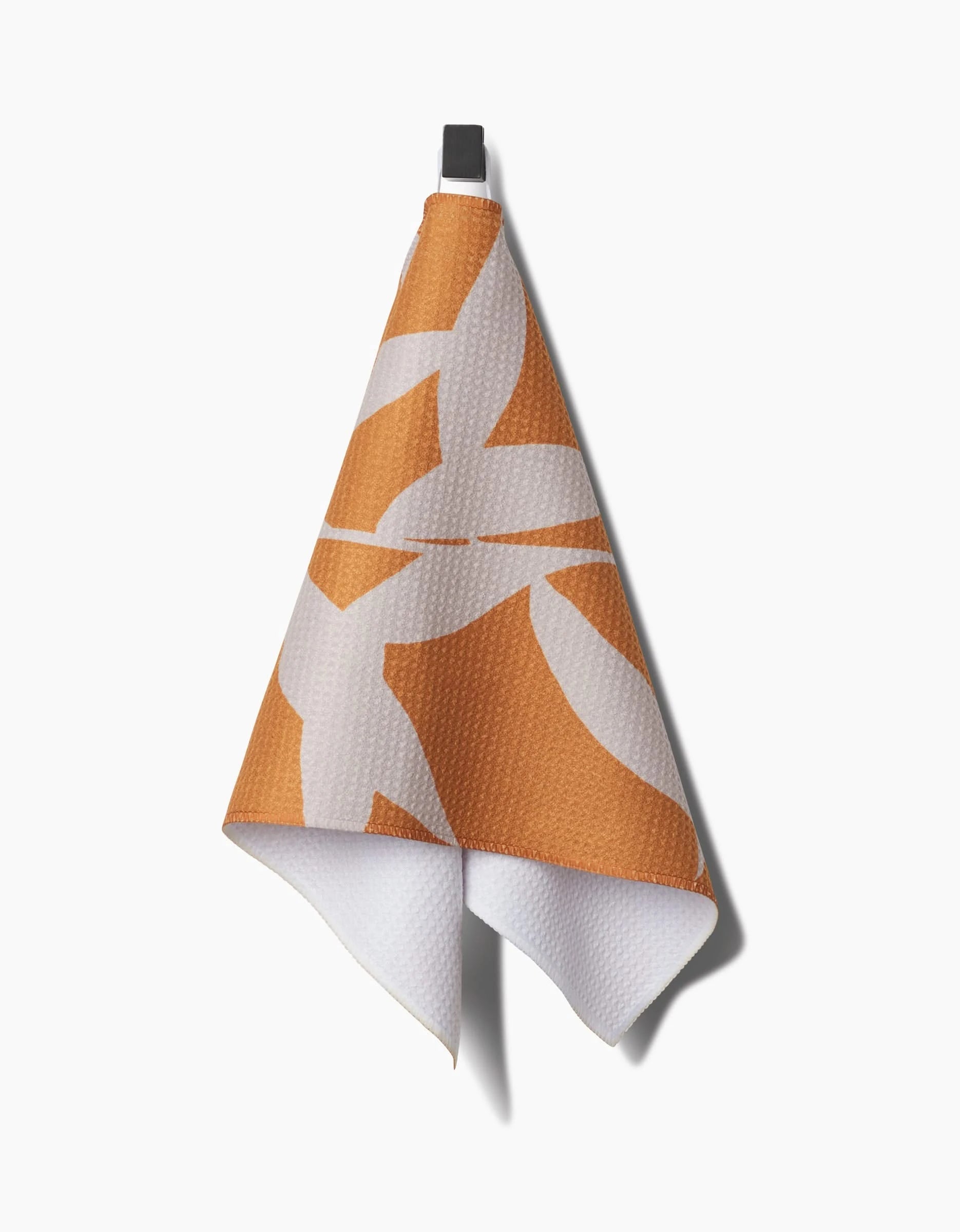 The Solidago bar towel by PrintedPlant, made from textured Geoweave fabric, showcases a geometric orange and white pattern. Hanging from a black hook against a white background, it brings modern style to any space.