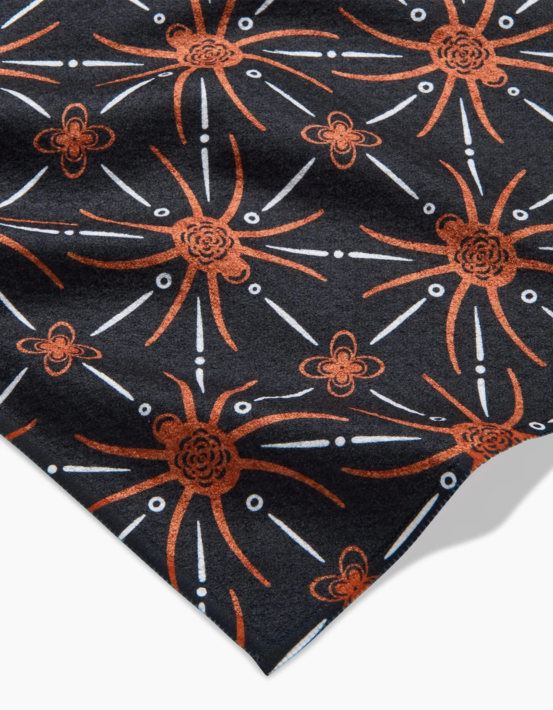 The Spider Geometric towel by Sarah Daugherty is black with an ultra-absorbent fabric, featuring an abstract orange and white floral starburst pattern with radiating lines, dots, and clusters. The fabric is shown folded at the bottom corner.