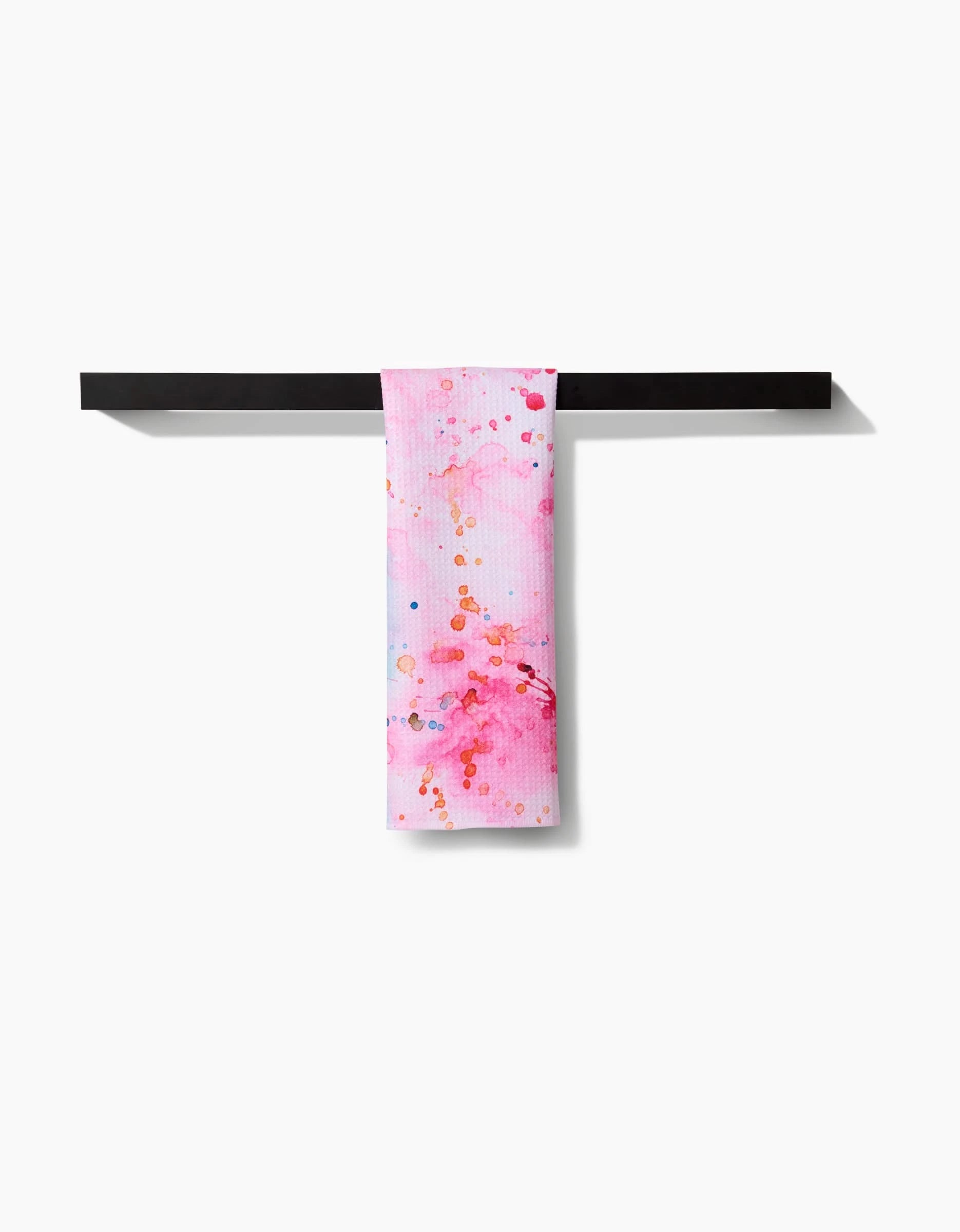 The Splash of Love ultra-absorbent towel by Sara Mari, featuring abstract pink, red, and blue watercolor splashes, hangs over a sleek black bar against a plain white background.