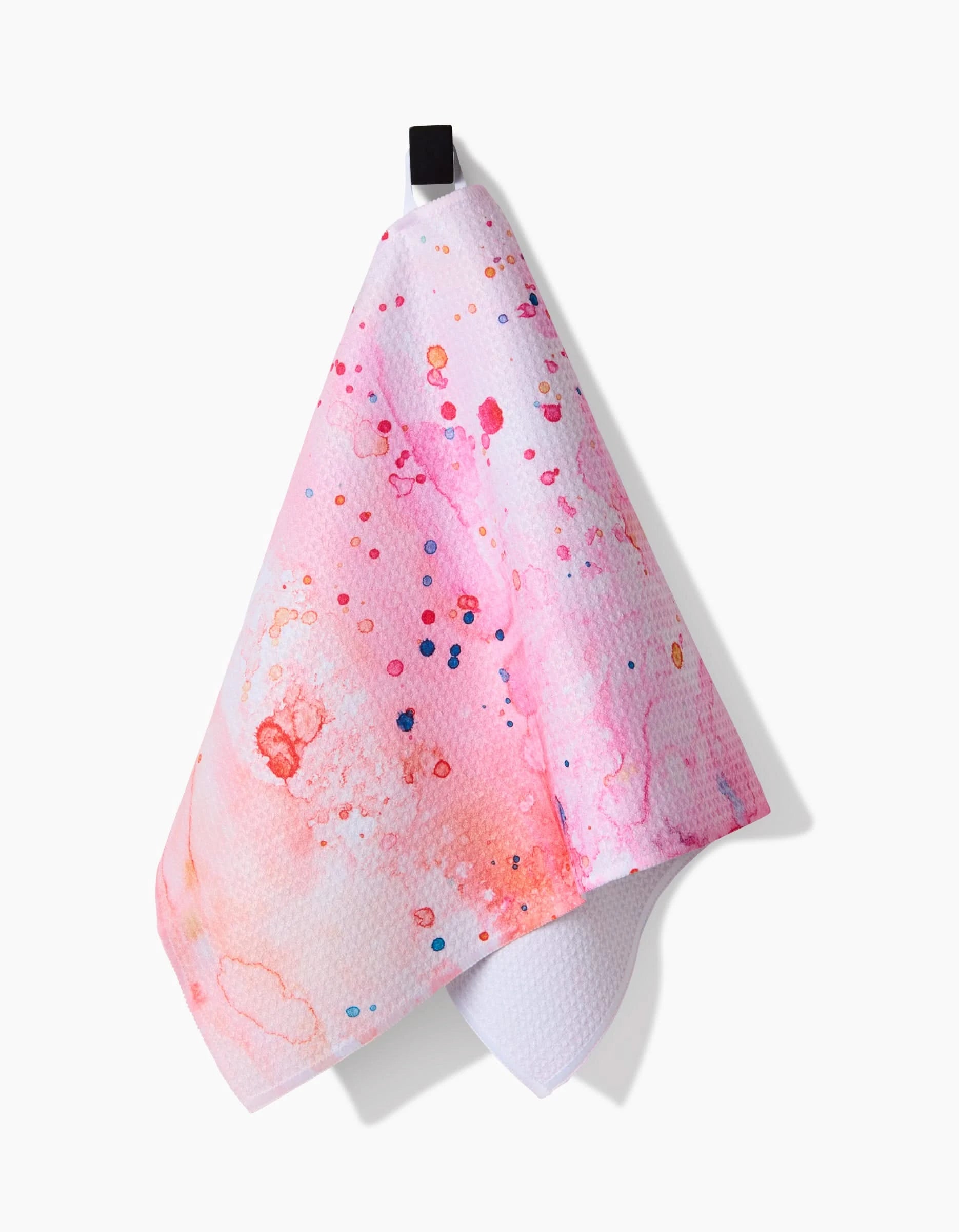 The Sara Mari Splash of Love white recycled kitchen towel features pink, orange, and red watercolor-style splatters and hangs from a black loop against a white background.