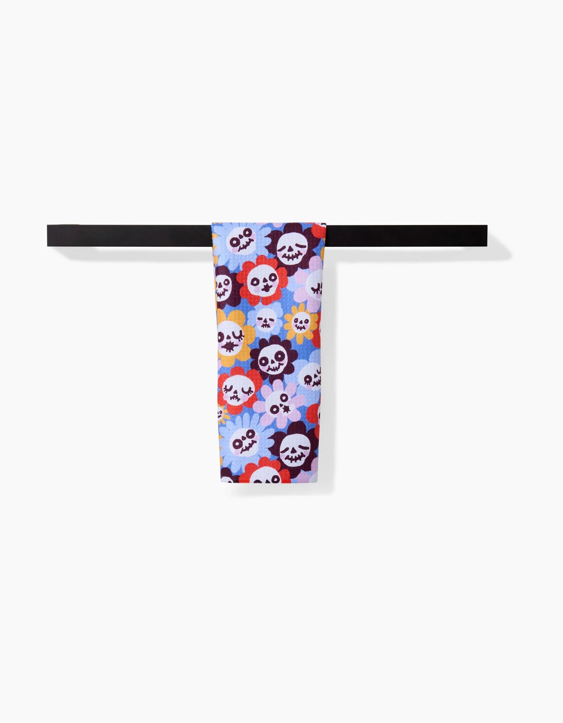 The So Lazo Spooky Bouquet towel, ultra-absorbent and colorful with cartoon skulls and flowers in red, purple, yellow, and white, hangs over a simple black rod against a white background.