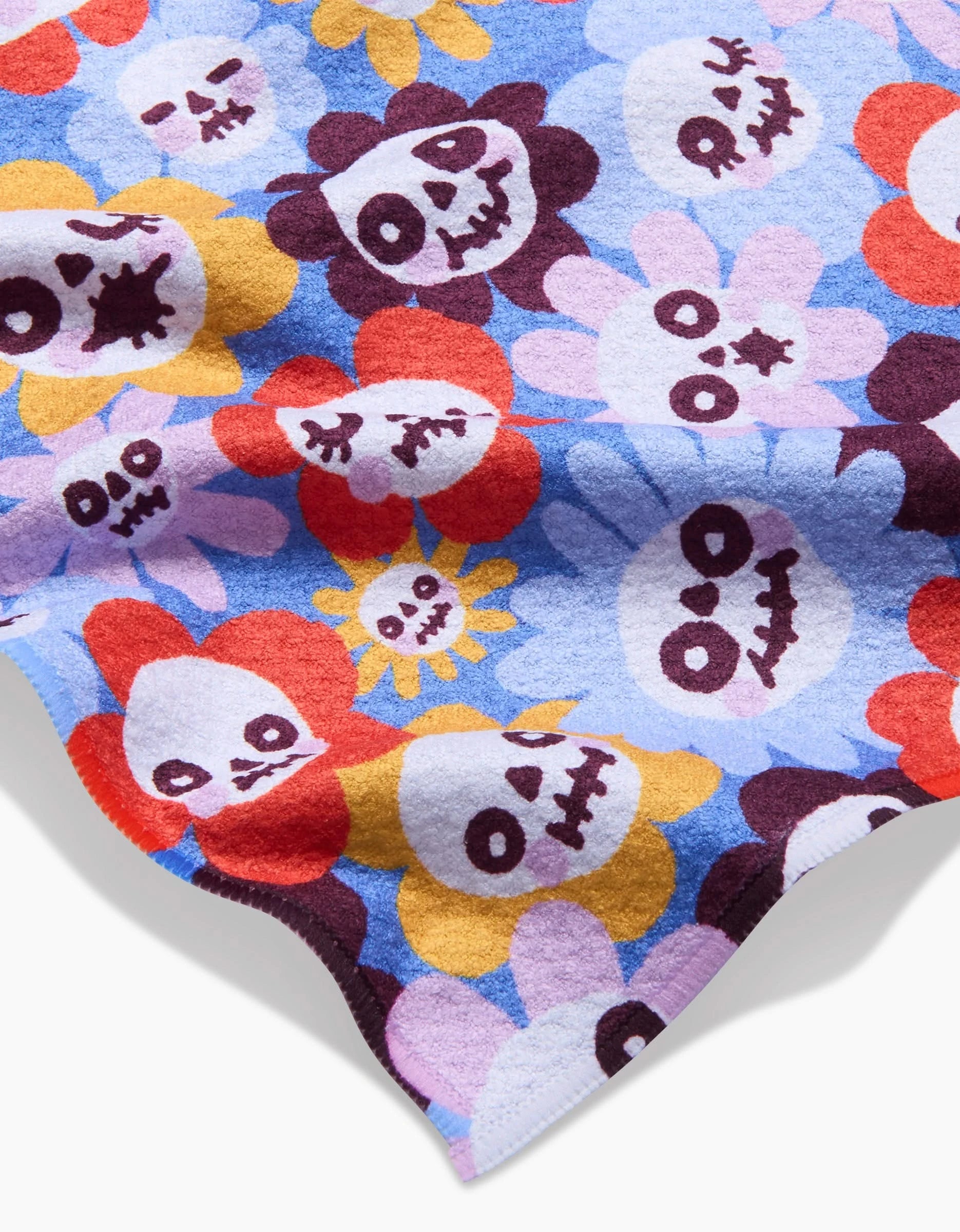 So Lazos Spooky Bouquet bar towel features cartoon flowers with smiling skull faces in red, yellow, purple, and white on a blue background. Made from ultra-absorbent recycled materials.