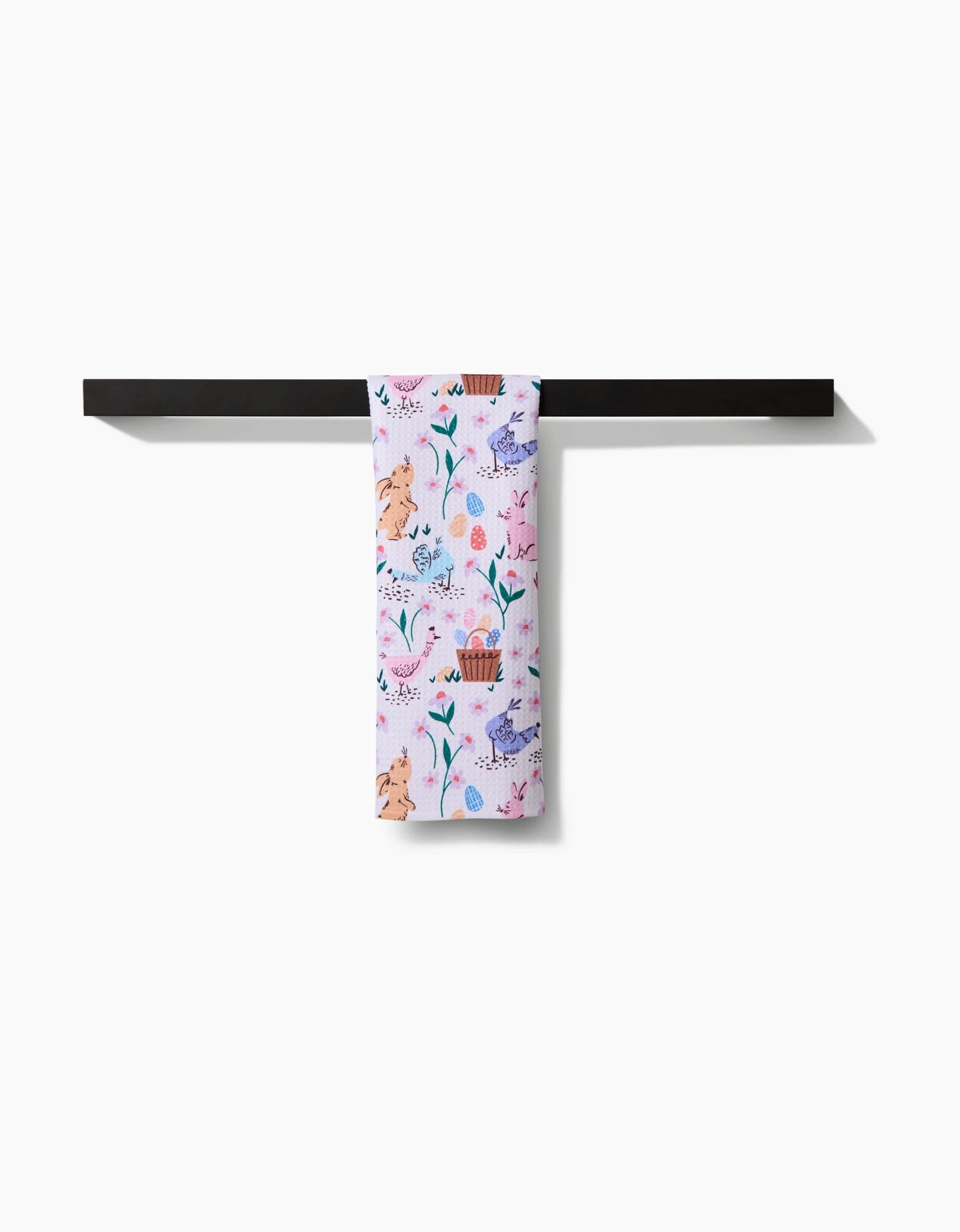 The “Spring Bunnies” towel by Charlie Rowan Designs, featuring pastel hues and whimsical animal and plant prints, hangs neatly on a black horizontal towel bar against a white backdrop.