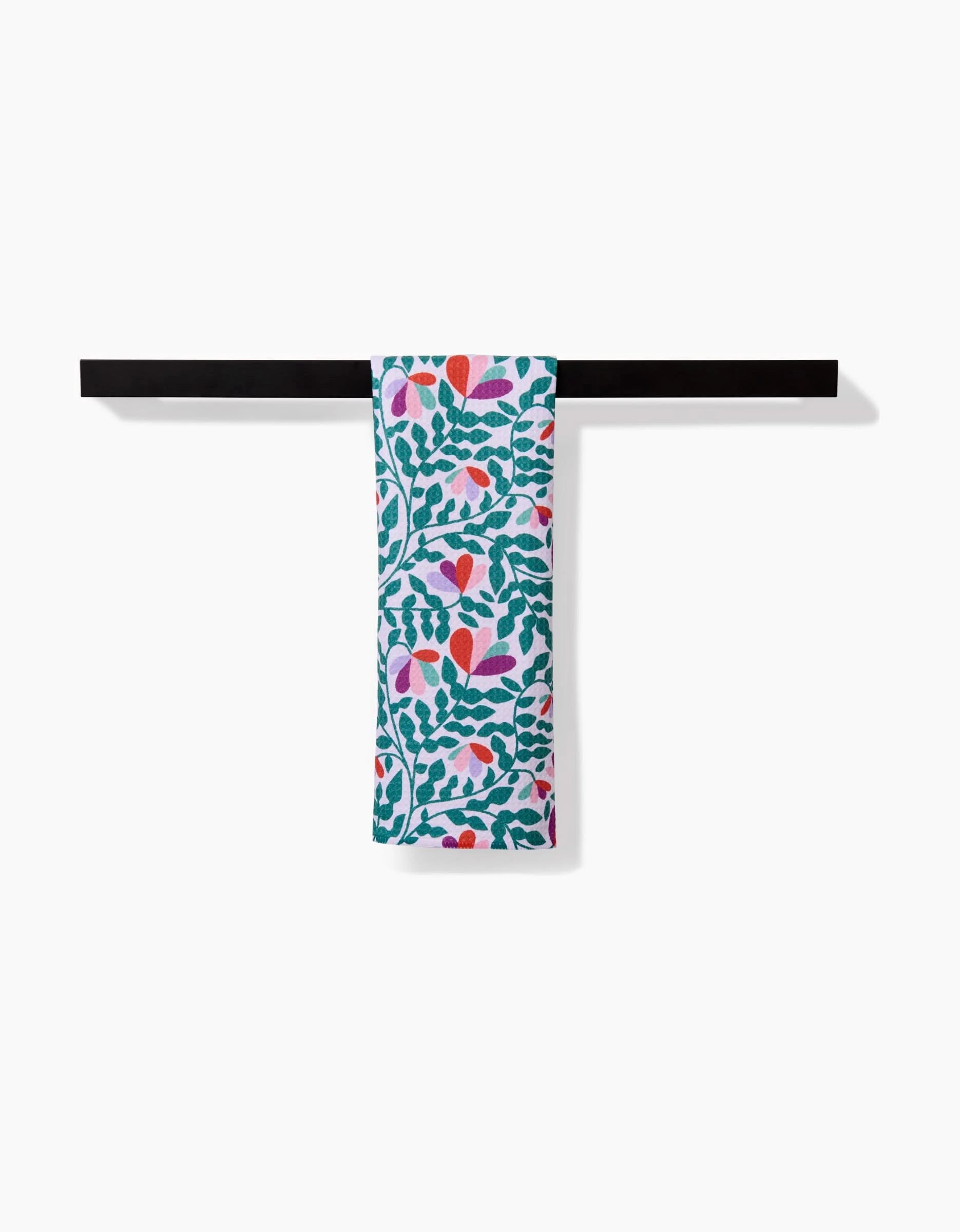 The Claire Iglesias Spring Wavy Leaves bar towel features a vibrant green, red, pink, and purple floral and leaf pattern. Made from soft, durable Geoweave fabric, it’s shown draped over a black bar against a white background.