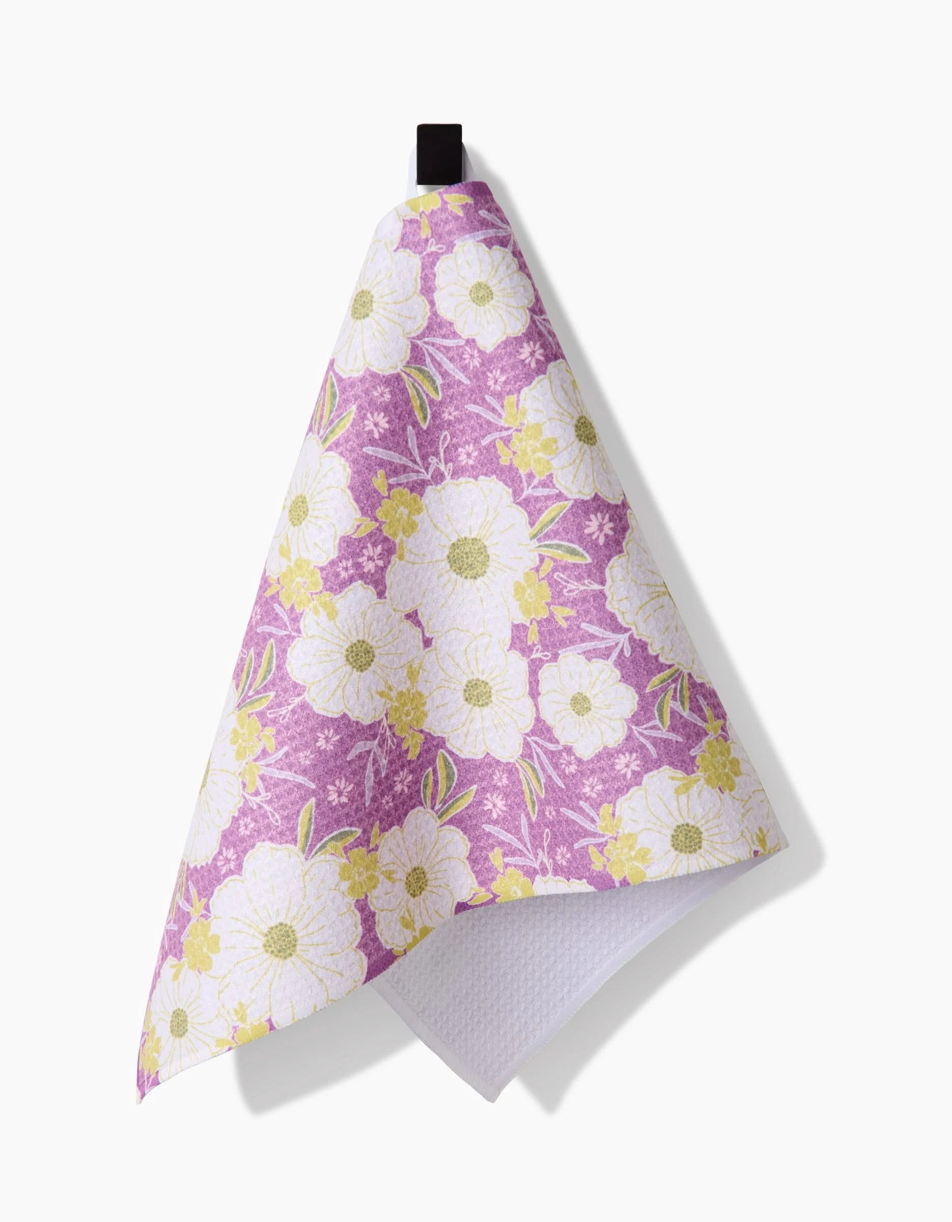 The Springtime Floral Bar Towel by Ashley Celena features a purple background with white and yellow flowers, made from recycled materials, and hangs neatly from a black loop.