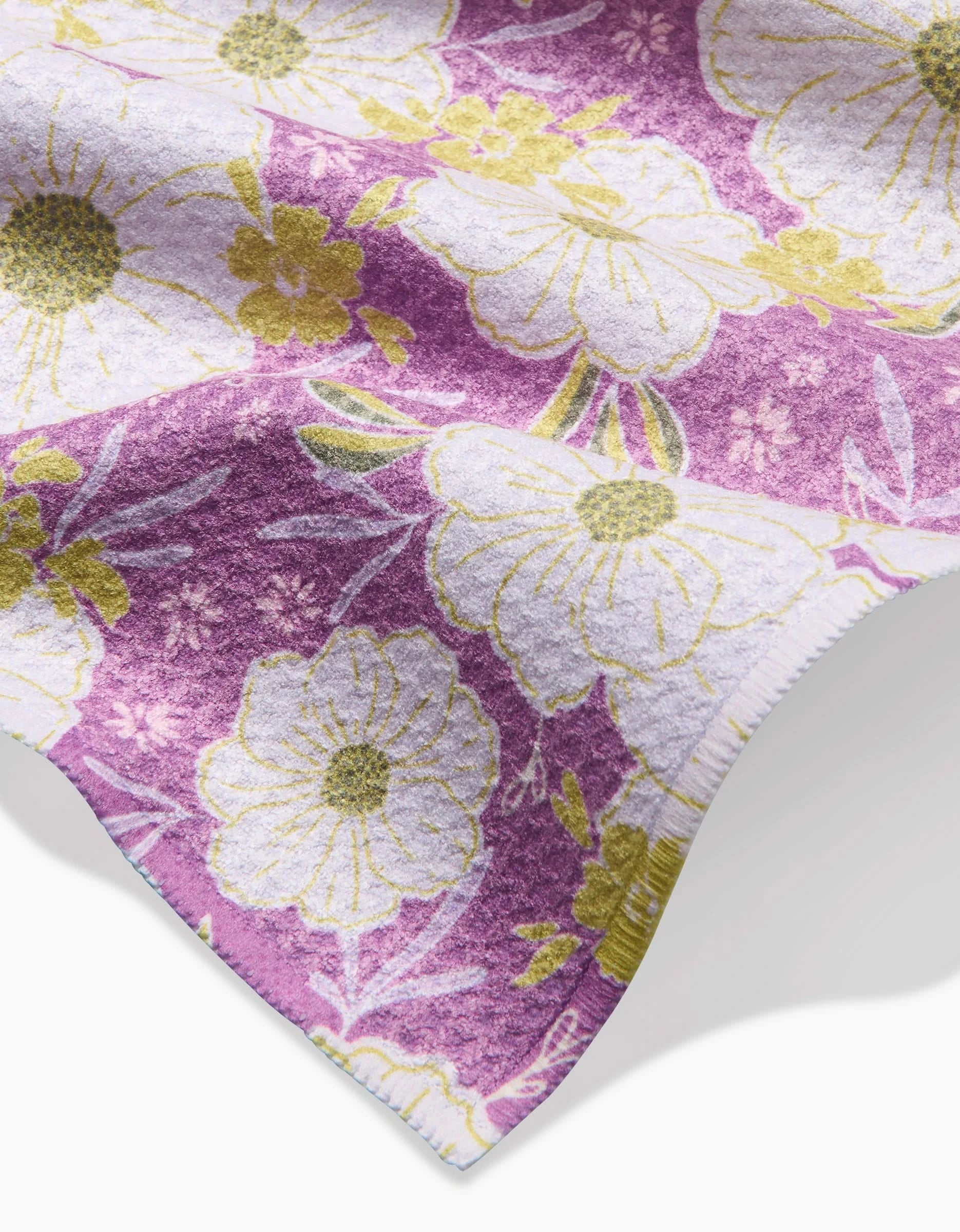 A close-up of the Ashley Celena Springtime Floral Bar Towel, crafted from Geoweave fabric with large white and yellow flowers plus green leaves on a purple background; the soft, textured material has curved edges.