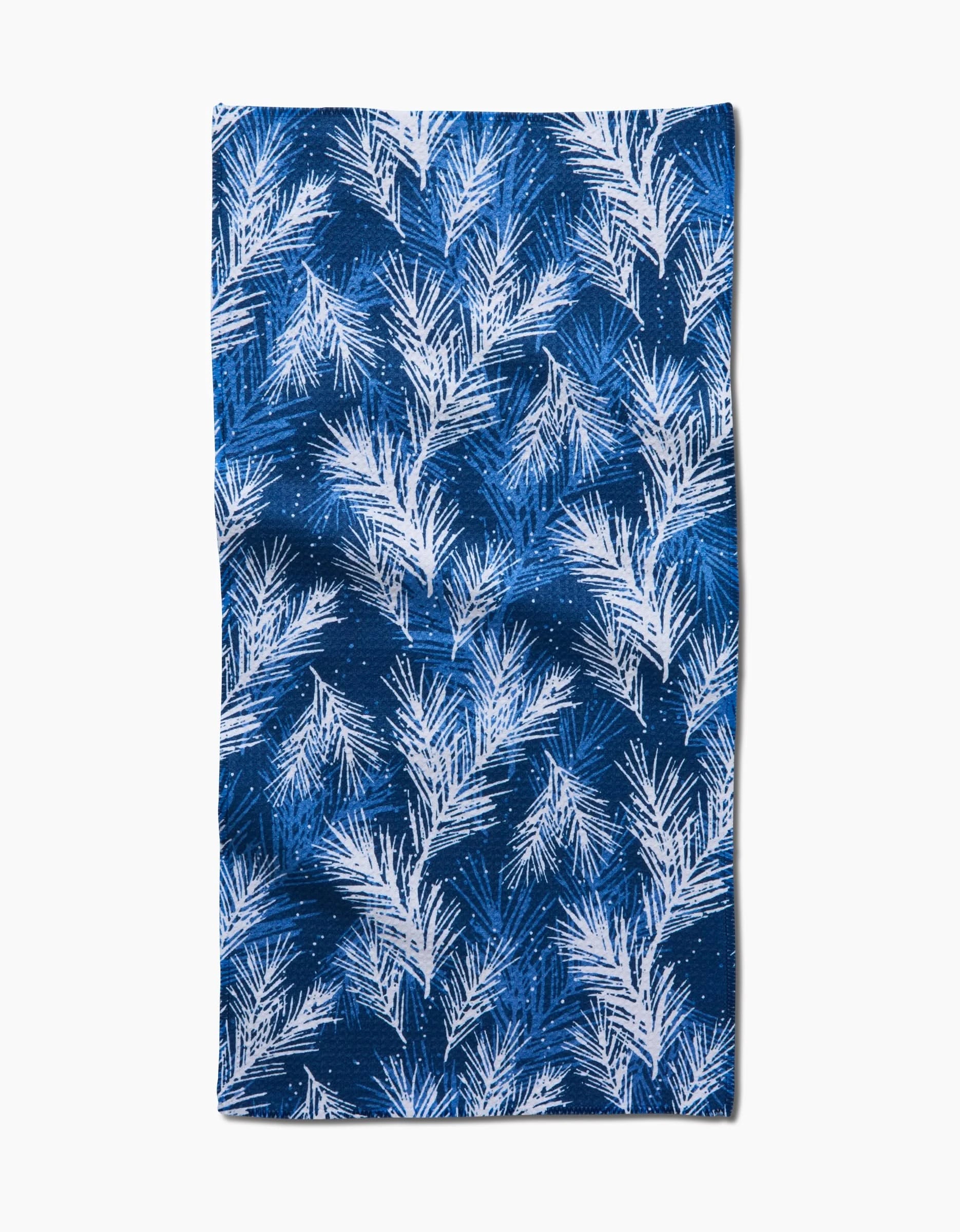 The Kris Ruff Sprinkled Spruce towel is rectangular and blue, featuring a dense white fern or palm leaf pattern in various directions over a gradient blue background for ultra-absorbency.