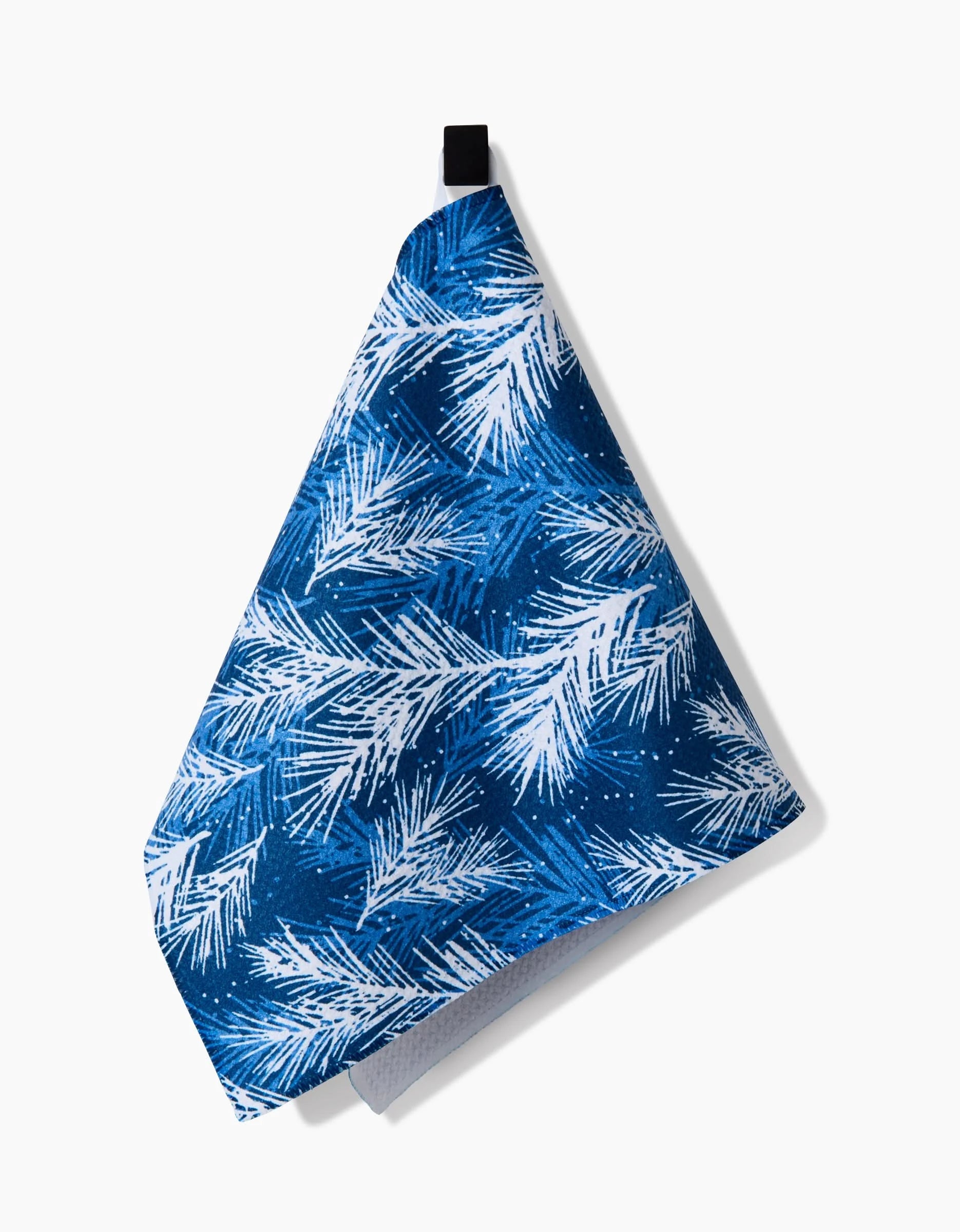 The Kris Ruff Sprinkled Spruce towel, featuring a white pine branch pattern on blue fabric, hangs by a black loop against a white background.