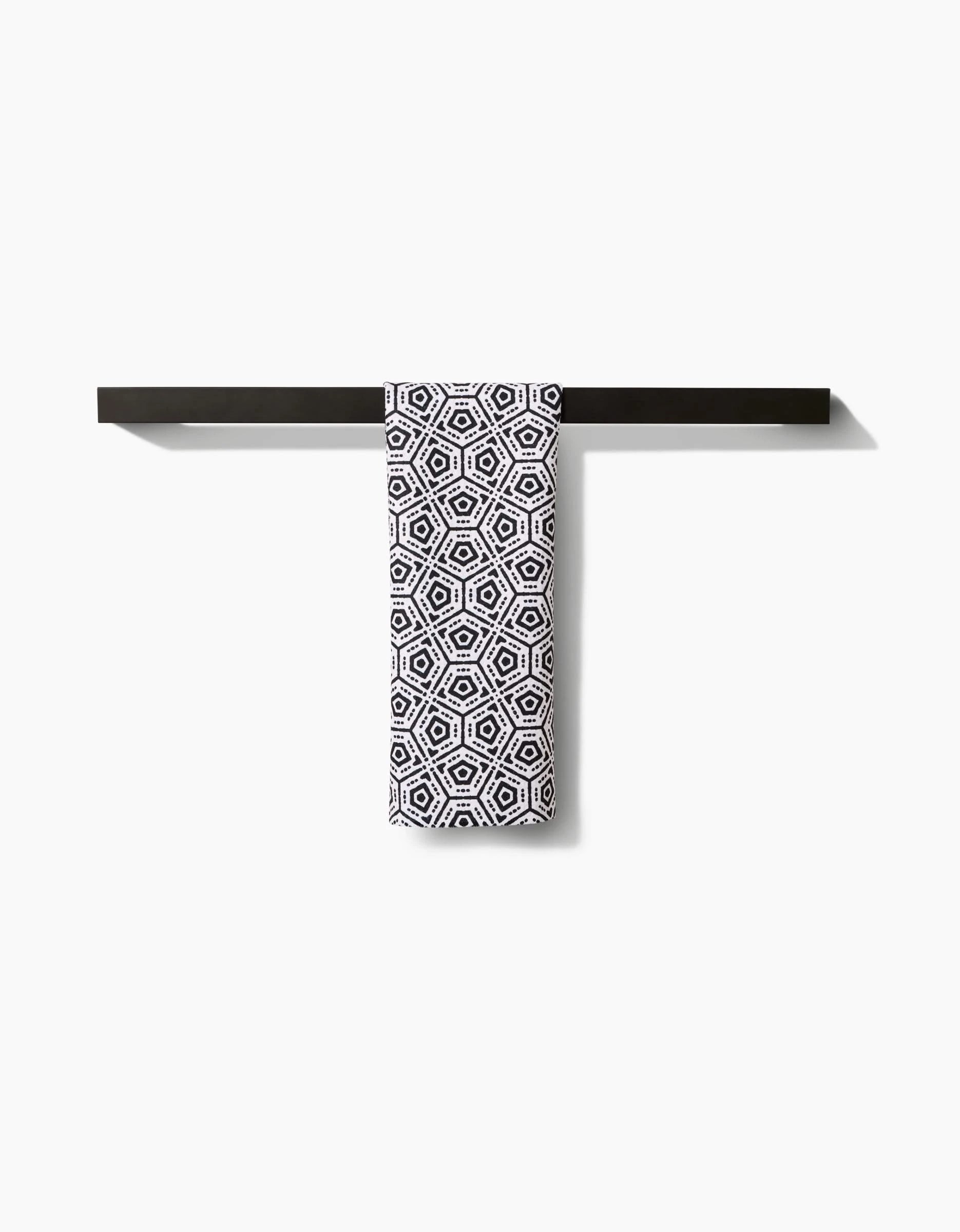 The Squeezebox Bar Towel by Katherine Berkeley, featuring a black-and-white geometric hexagonal pattern in Geoweave™ fabric, hangs neatly from a black towel bar against a plain white background.