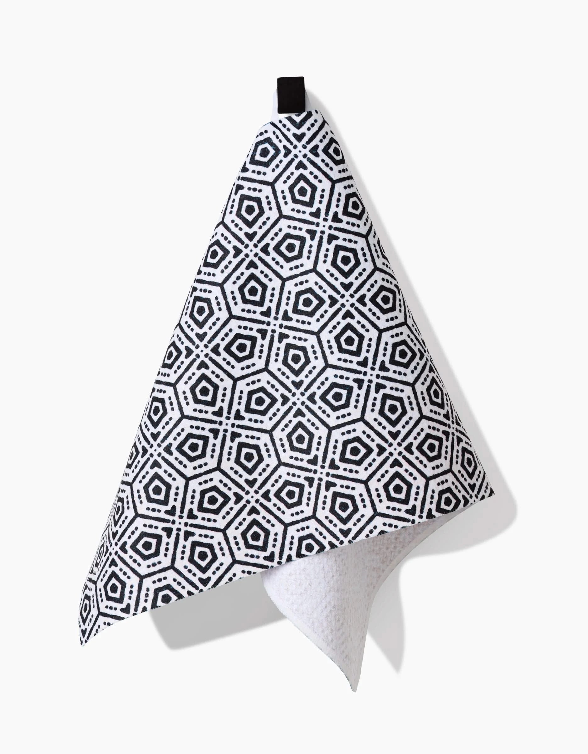 The Katherine Berkeley Squeezebox Bar Towel, featuring a black and white geometric hexagonal design, hangs from a black loop against a white background. Made from Geoweave™ fabric, it casts a shadow to the right.