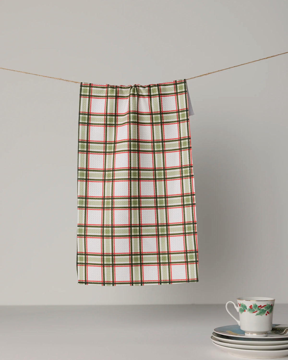 A Stocking Plaid tea towel by Geometry, made from recycled materials and featuring red, green, and white stripes, hangs on a string above a stack of plates and a teacup with floral patterns on a plain surface.