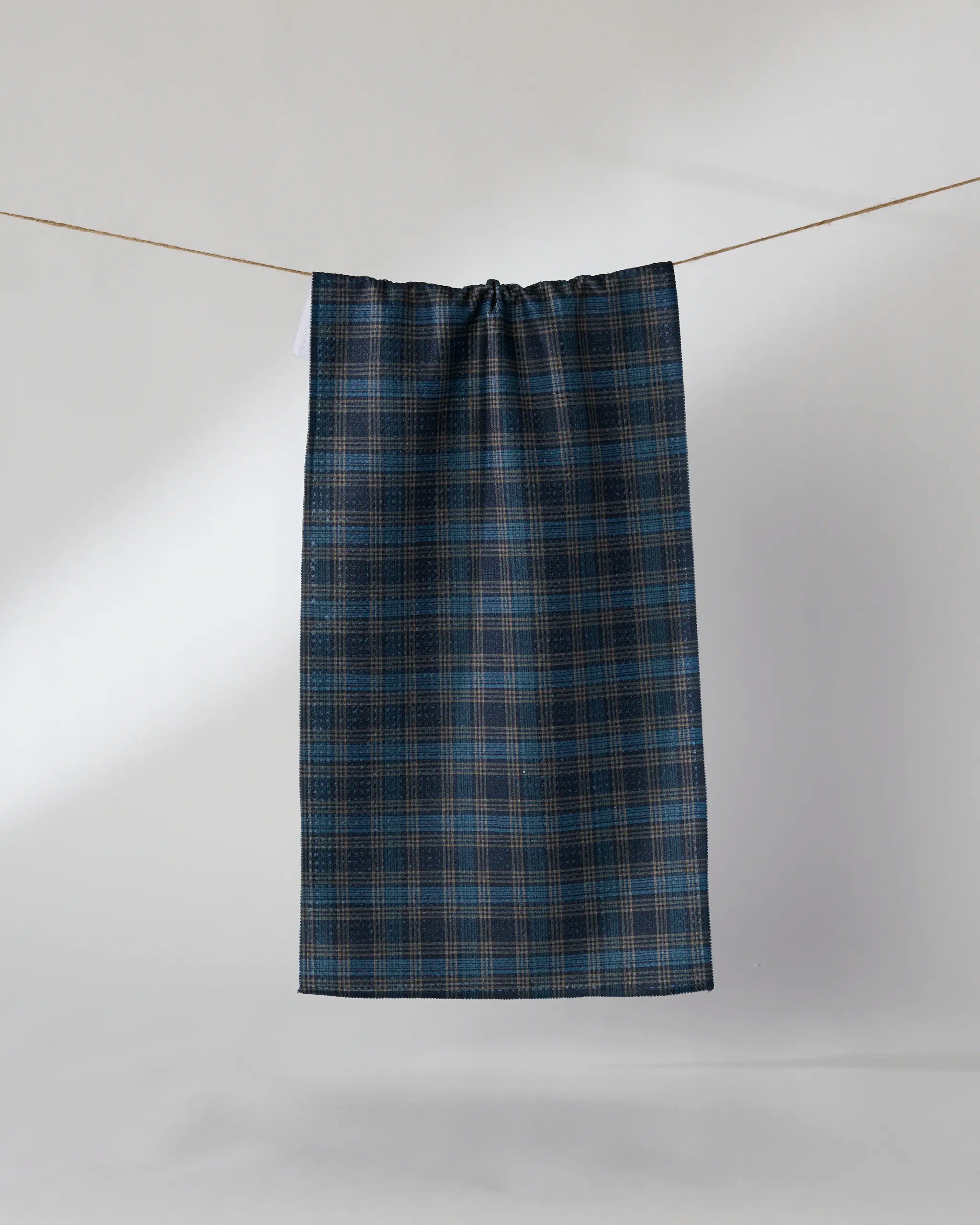 The Geometry Stonegate bar towel, made from recycled materials, in a dark blue and brown plaid pattern, is displayed hanging on a clothesline against a plain light gray background.