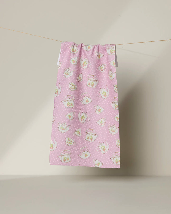 Geometry’s “Strawberry Tea” recycled tea towel in pink, adorned with white polka dots and whimsical teapot and teacup illustrations, hangs on a clothesline against a neutral background.