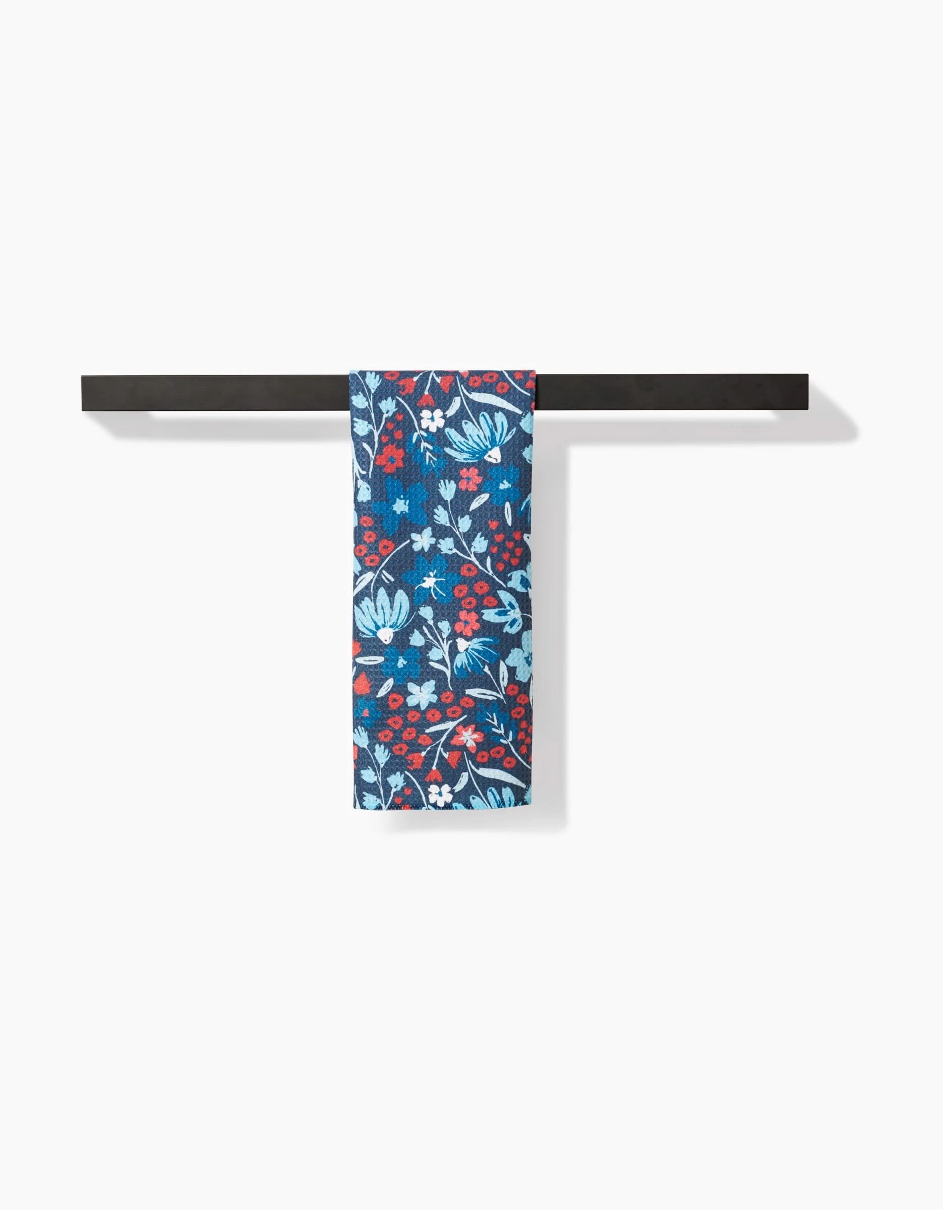 The Summer Bloom scarf by Hufton Studio, made from recycled materials, showcases a red, white, and light blue floral pattern on blue and is elegantly draped over a black rod against a white background.