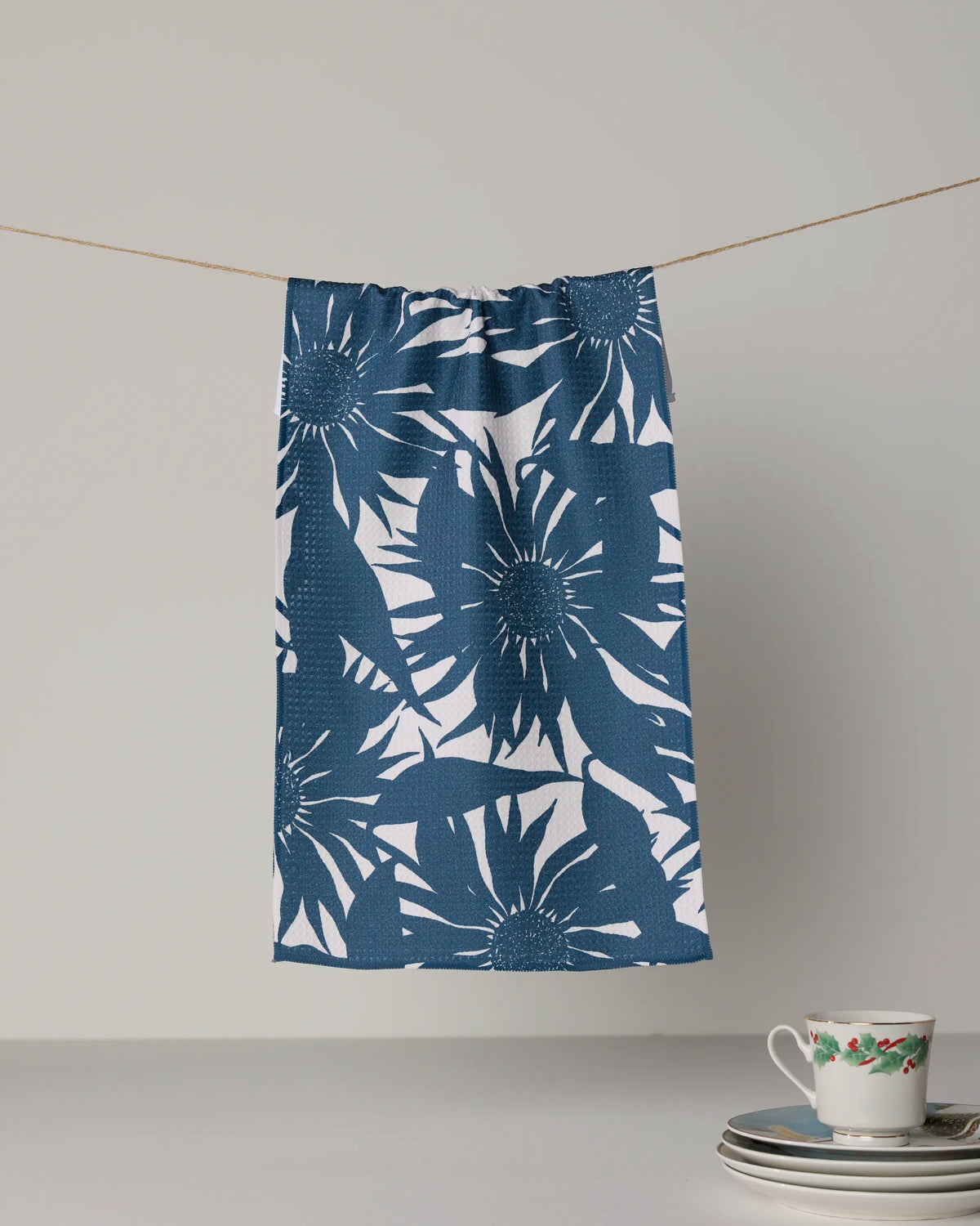 The Geometry Sundara Silhouette Frost tea towel, crafted from recycled materials with a bold blue and white floral design, hangs on a string above a table set with stacked plates and a floral teacup.