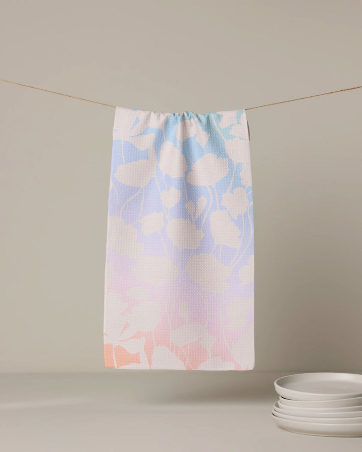 The Geometry Sunlit Silhouette kitchen towel, featuring a pastel leaf pattern and eco-friendly design, hangs above a stack of white plates against a neutral background.