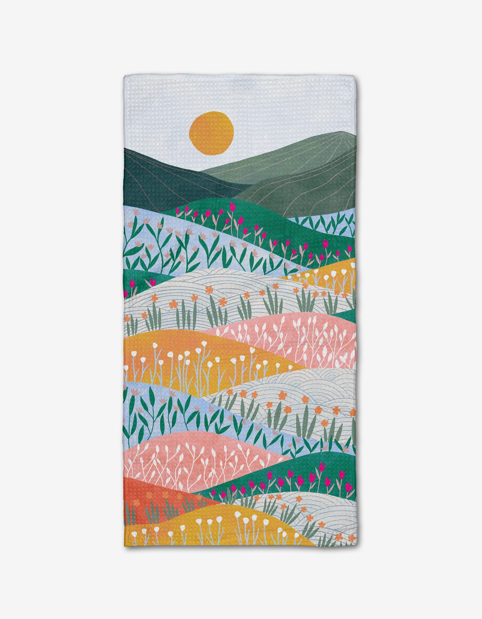 The Sunrise Mountains tea towel by Ceyda Alasar is made from recycled materials and features a vibrant landscape with rolling hills, pink, orange, and green flowers, plus a yellow sun against a blue sky.
