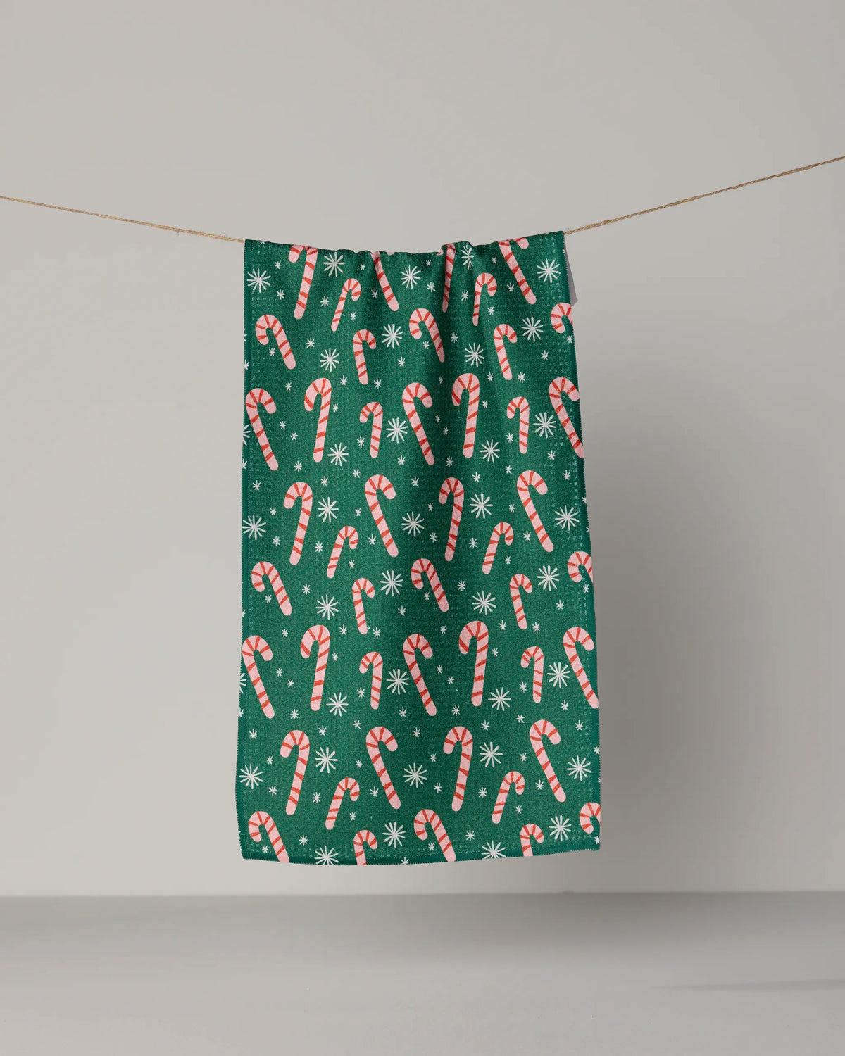 The Geometry Sweet Candy mini towel, featuring a festive green design adorned with red and white candy canes and white snowflakes, is crafted from recycled materials and hangs on a clothesline against a plain background.