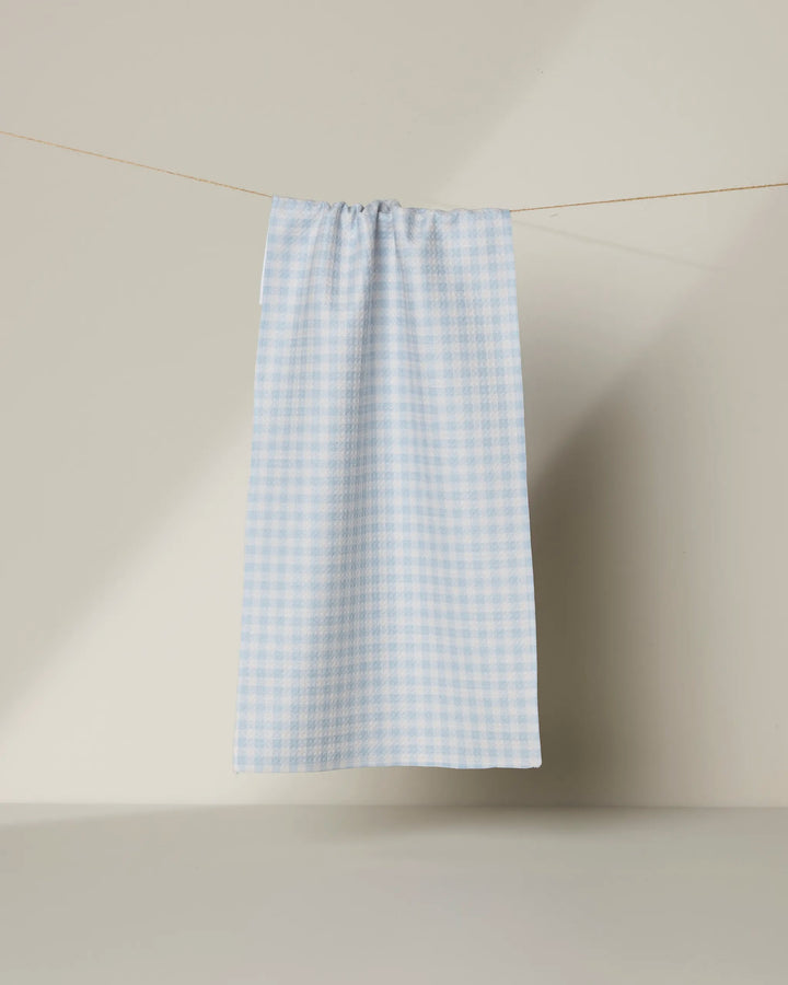The Geometry Sweet McKelle towel, featuring a light blue and white gingham pattern, hangs from a string against a plain beige background.