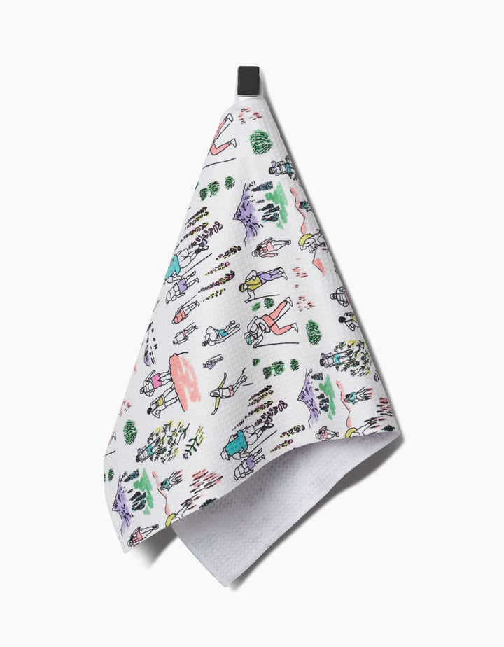 The Elisabeth Wing Take A Hike bar towel features colorful, playful drawings of hikers, trees, mountains, and animals on ultra-absorbent Geoweave fabric, with a black hanging loop on a white background.