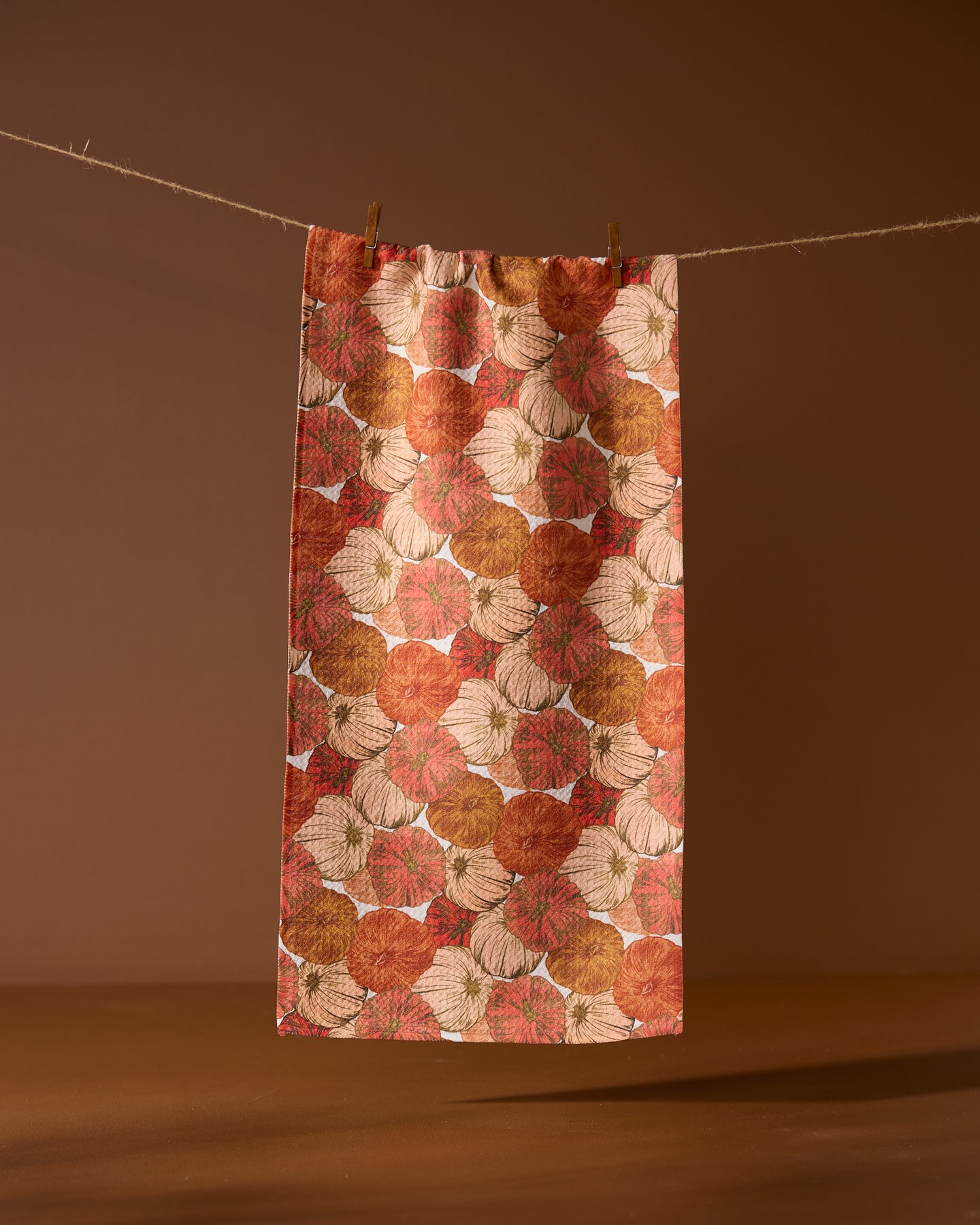 The Geometry Tendril towel, featuring an ultra-absorbent design with orange, brown, and beige pumpkin patterns, hangs on a string against a plain brown background, held by two wooden clothespins.