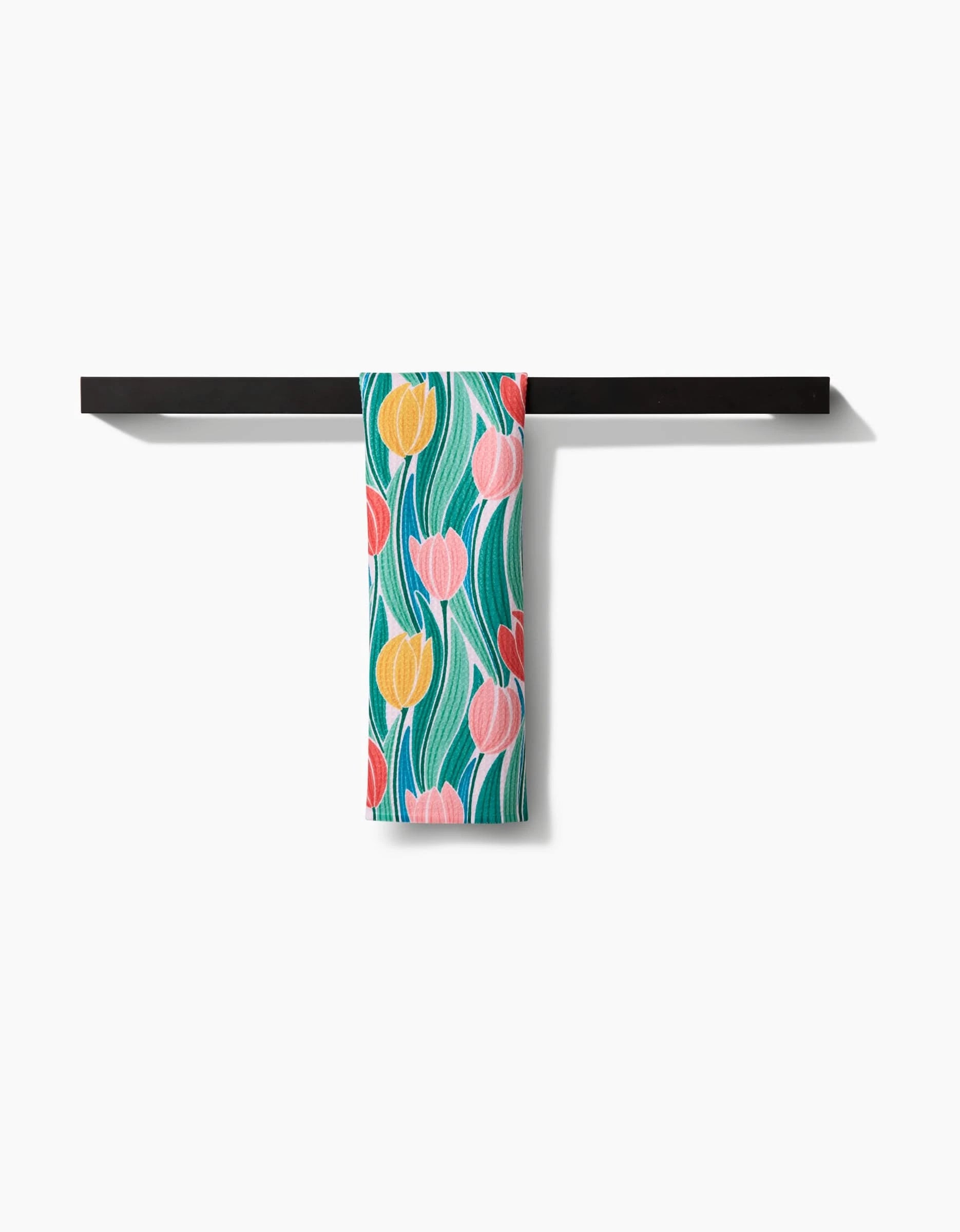 The Tranquil Tulips bar towel by Sarah Daugherty, crafted from recycled materials and featuring a vibrant tulip pattern in yellow, pink, and red with green leaves, hangs over a black towel bar against a white background.