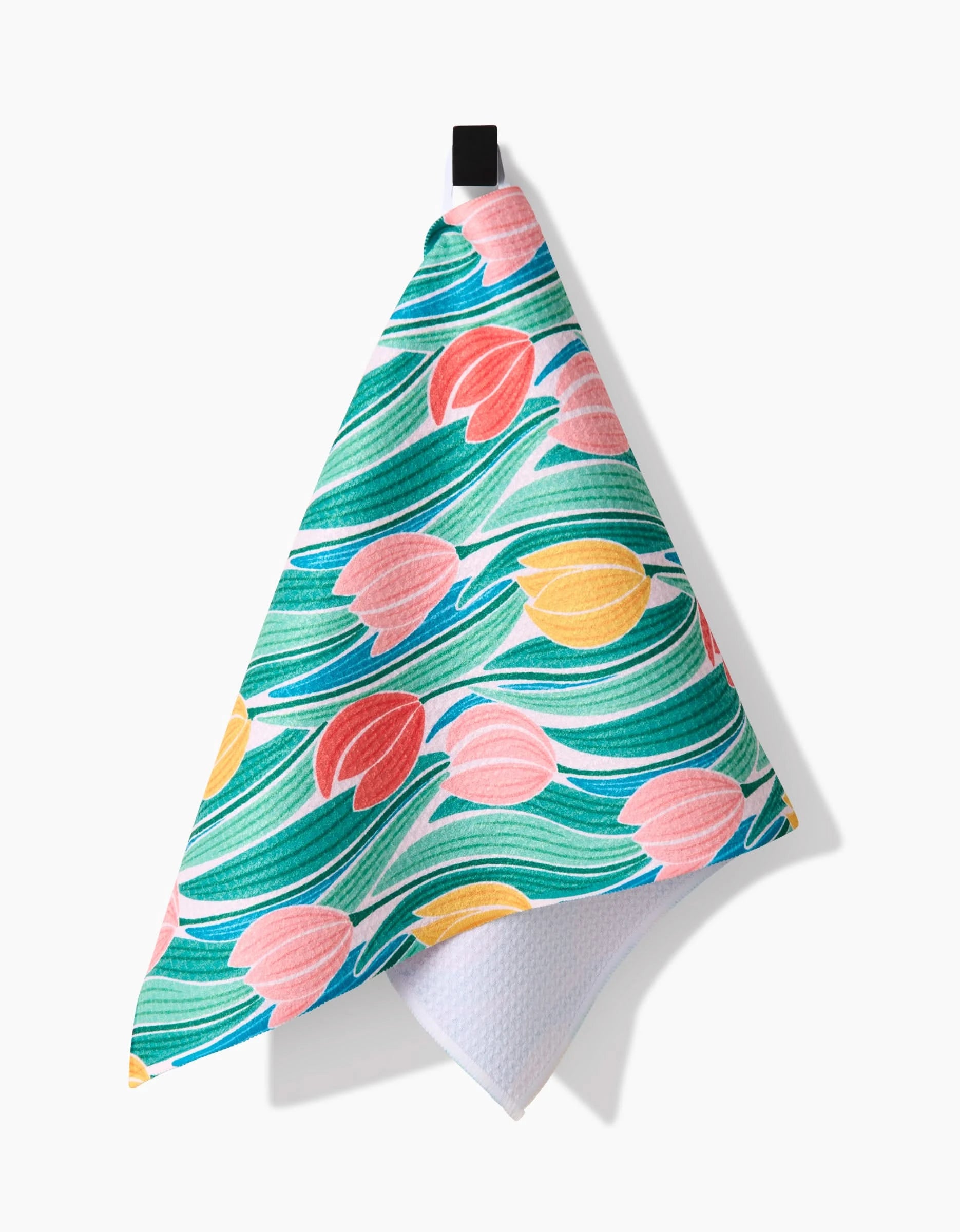The Tranquil Tulips bar towel by Sarah Daugherty is crafted from recycled materials and showcases a vibrant pattern of pink, yellow, and red tulips with green leaves, hanging from a black loop against a white background.