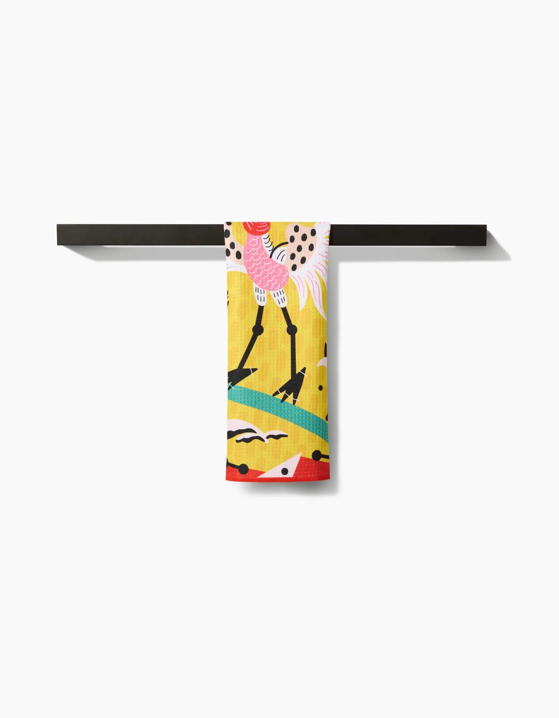 The Tuiuiú bar towel by Camila Leão features a whimsical pink and white bird on yellow, draped over a black bar for a cheerful look. Made from ultra-absorbent Geoweave fabric, it combines playful style with top performance.