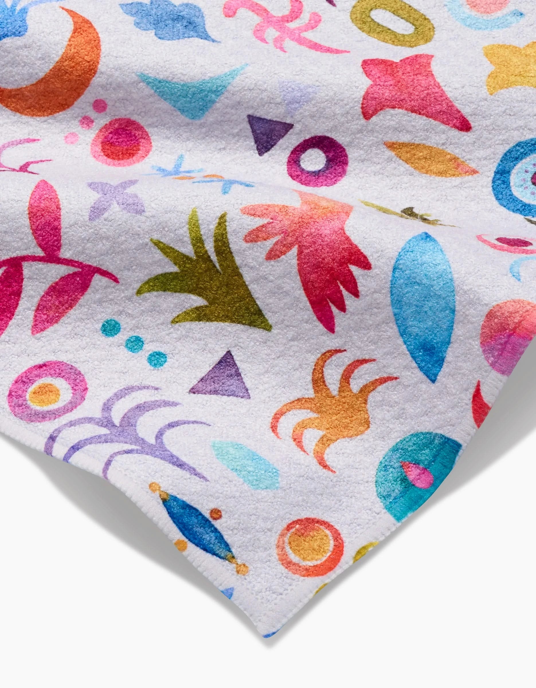 The Tula bar towel by Maayan Alper-Swan is made of white Geoweave fabric and features vibrant abstract patterns in pink, blue, orange, green, and purple with triangles, circles, and organic shapes scattered across the surface.