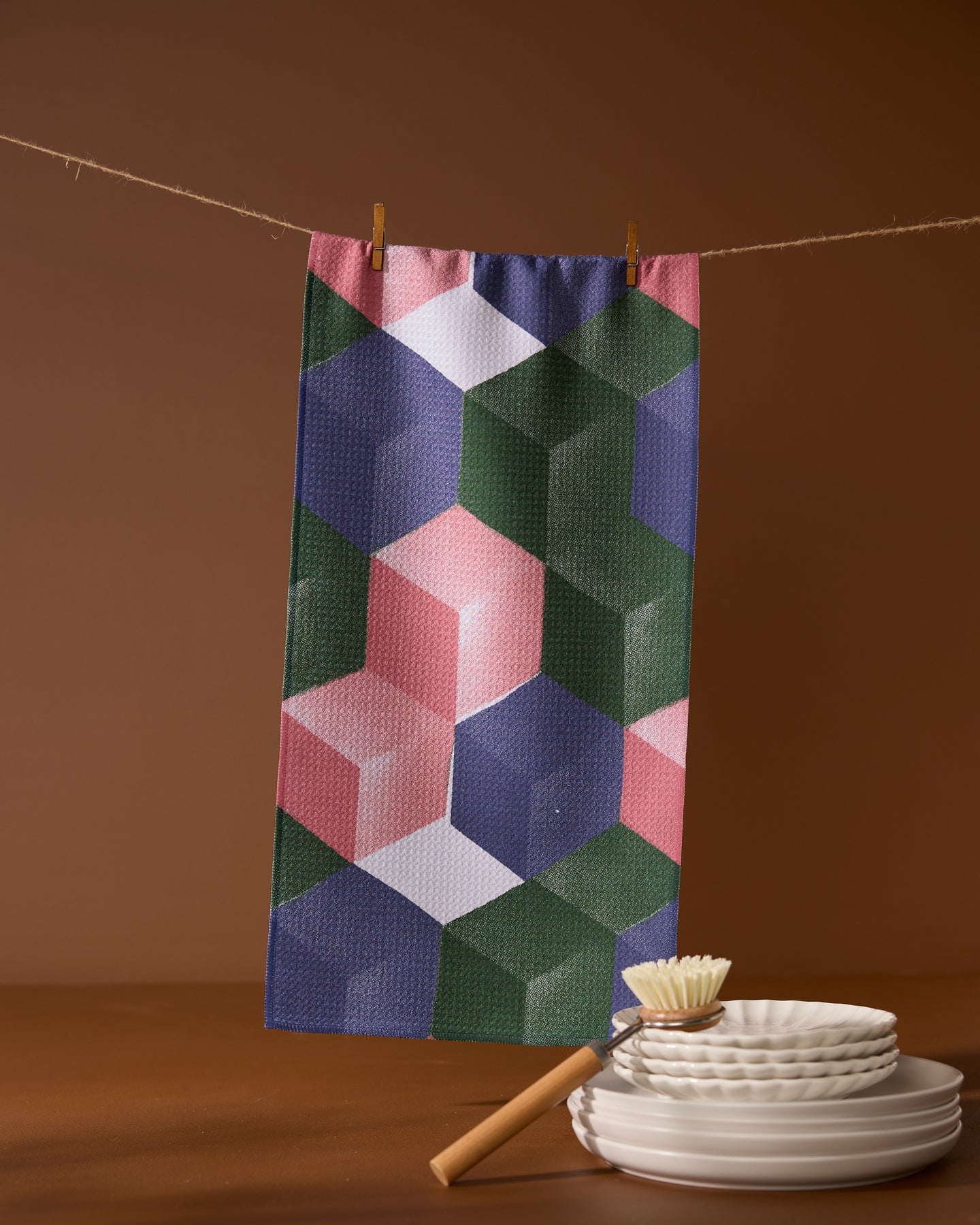 The Geometry Tumble ultra-absorbent towel with a colorful geometric pattern hangs on a clothesline with clothespins above stacked white plates and a dish brush against a brown background.