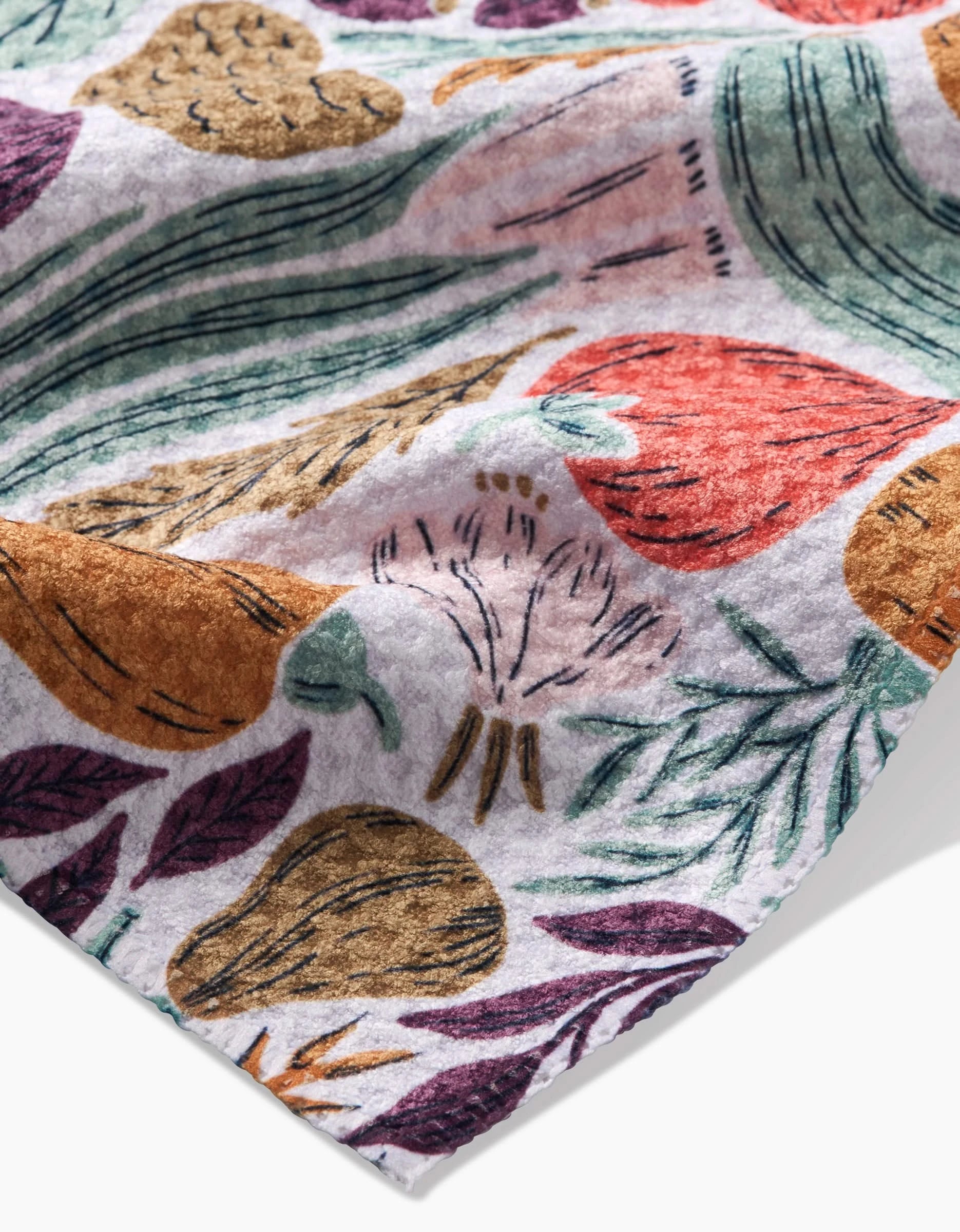 A close-up of Twila, a white bar towel by Amy MacCready, made from recycled materials with colorful vegetable and plant illustrations—carrots, radishes, onions, and leafy greens. Texture and vibrant patterns are prominently featured.