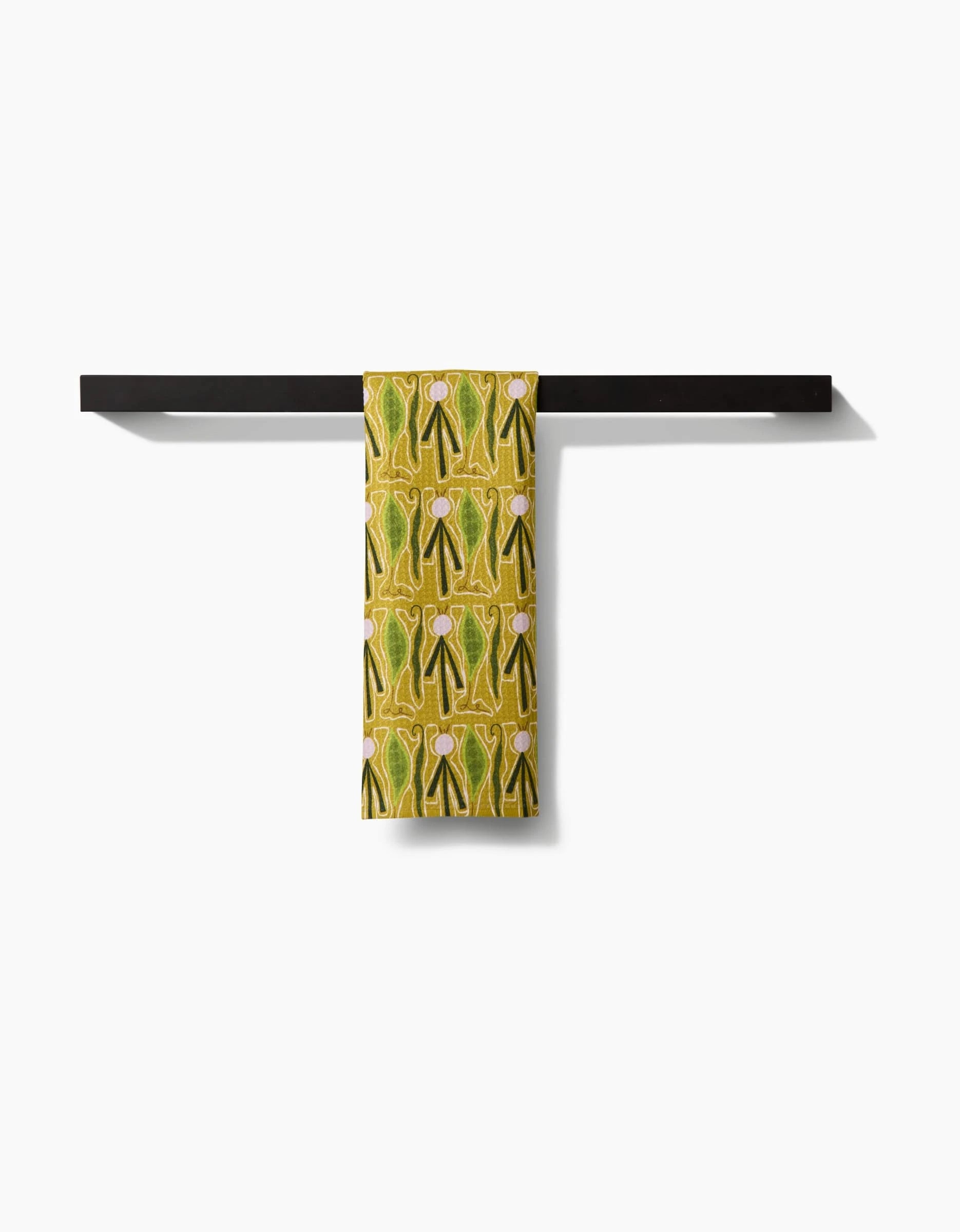 The Veg on Parade bar towel by Lizzie Clark, featuring a yellow-green pattern and made from recycled materials, is neatly draped over a minimalist black bar with a plain white backdrop.