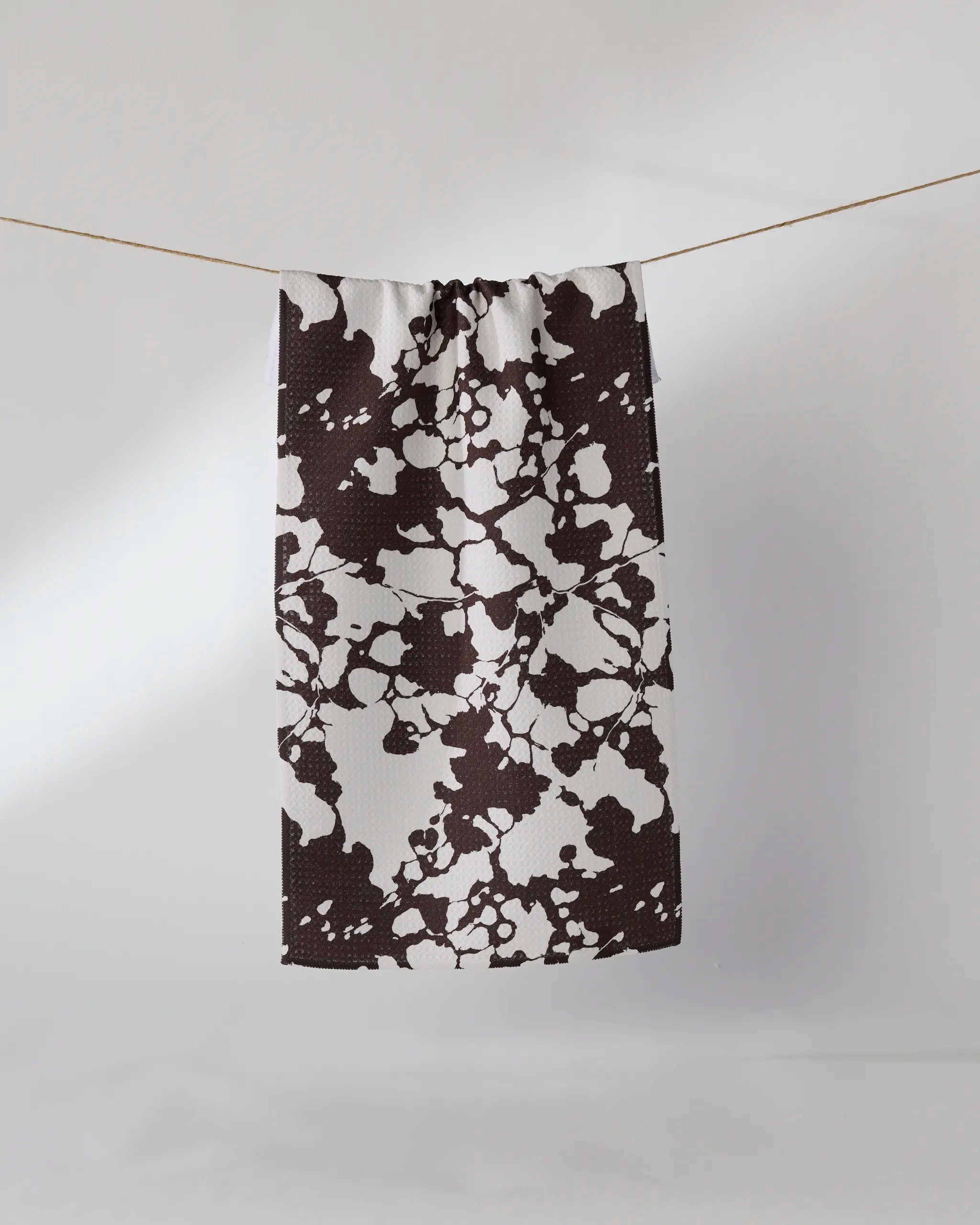 The Geometry Vine Noir rectangular bar towel, crafted from recycled materials, displays a black and white abstract splatter pattern and hangs on a clothesline against a plain white background.