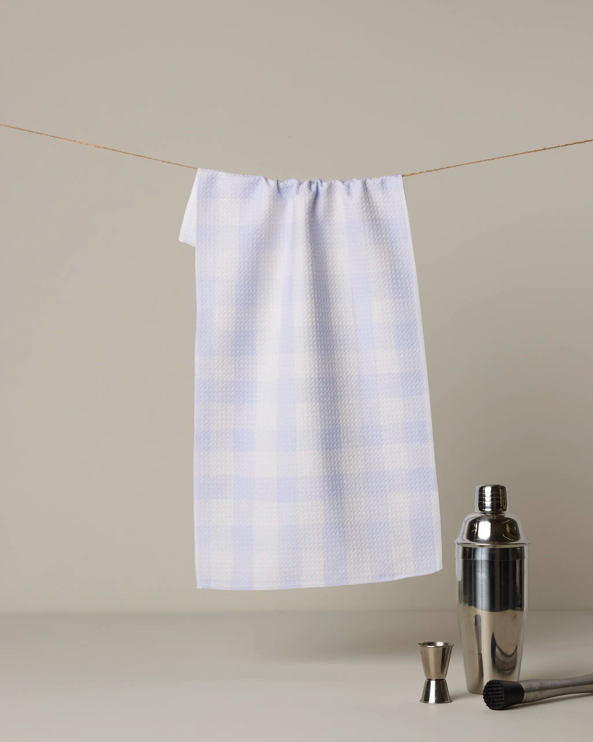 The Geometry Violetta bar towel, featuring a white and light blue checkered pattern, hangs on a clothesline with a cocktail shaker, jigger, and muddler arranged below against a plain background.