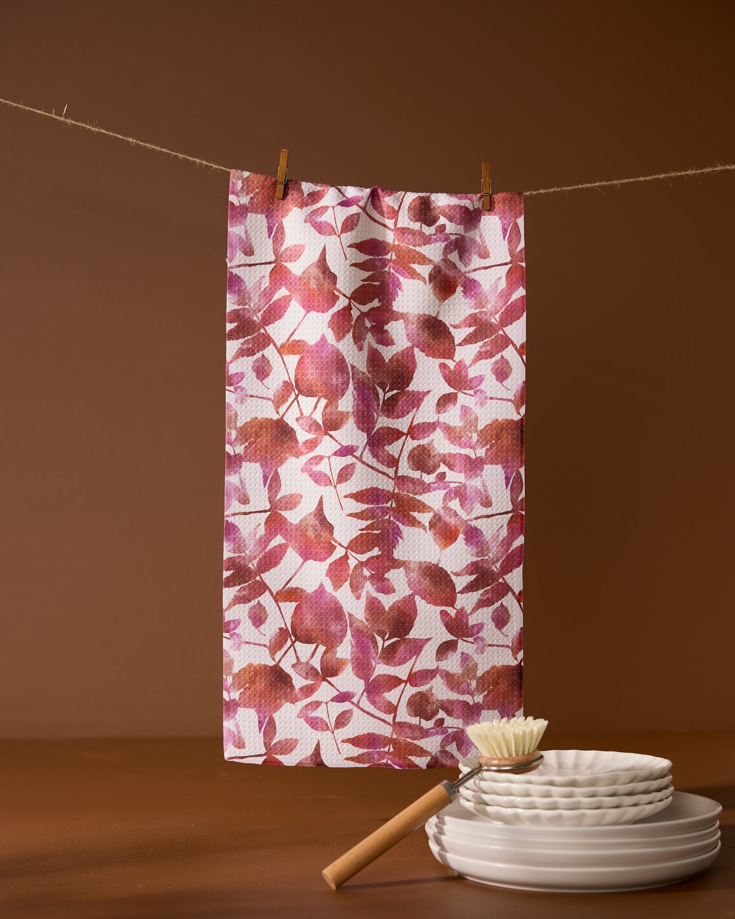 Geometrys Watercolor Dreaming recycled kitchen towel, featuring a red and pink leafy pattern, hangs on a clothesline above stacked white plates and a dish brush, all set against a coordinating brown background.
